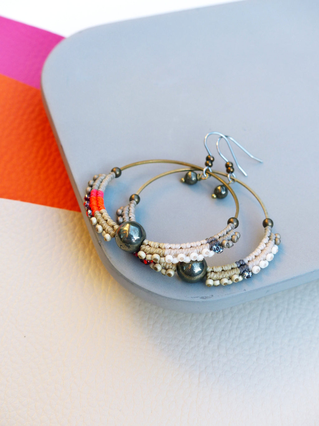 Peut inclure: Une paire de boucles d'oreilles créoles de style bohème. Les boucles d'oreilles sont composées d'un cerceau en métal doré, orné de fil beige et blanc tissé, de petites perles et d'une grosse perle gris foncé. Des touches de fil rouge et rose fluo ajoutent de la couleur.
