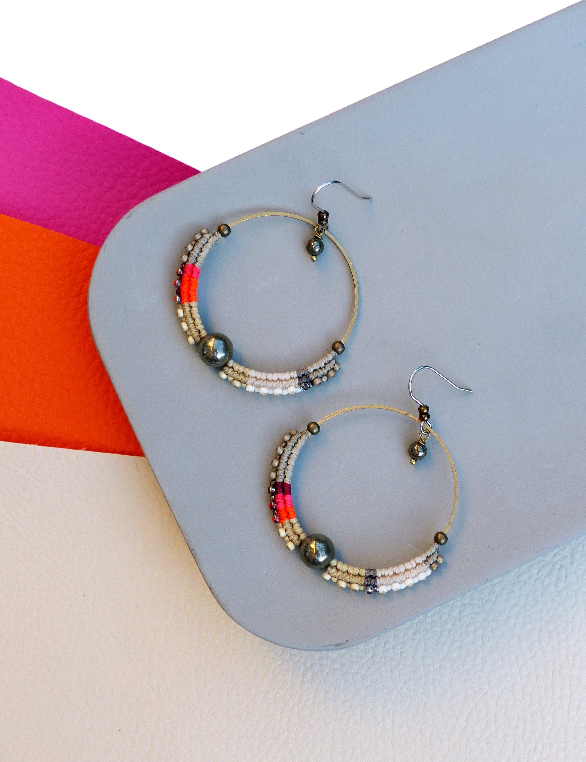 Peut inclure: Paire de boucles d'oreilles créoles avec une monture en métal doré. La moitié inférieure de chaque boucle est ornée d'un motif de petites perles blanches, grises, rouges et noires. Une grosse perle gris foncé est centrée sur chaque boucle.