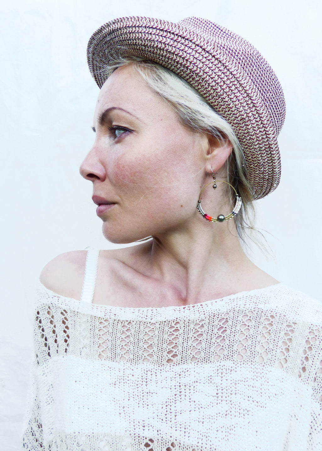 Peut inclure: Une femme porte un chapeau tissé avec un motif marron et beige et de grandes boucles d'oreilles créoles. Les boucles d'oreilles ont des perles colorées. Elle porte un pull en tricot ajouré blanc cassé.