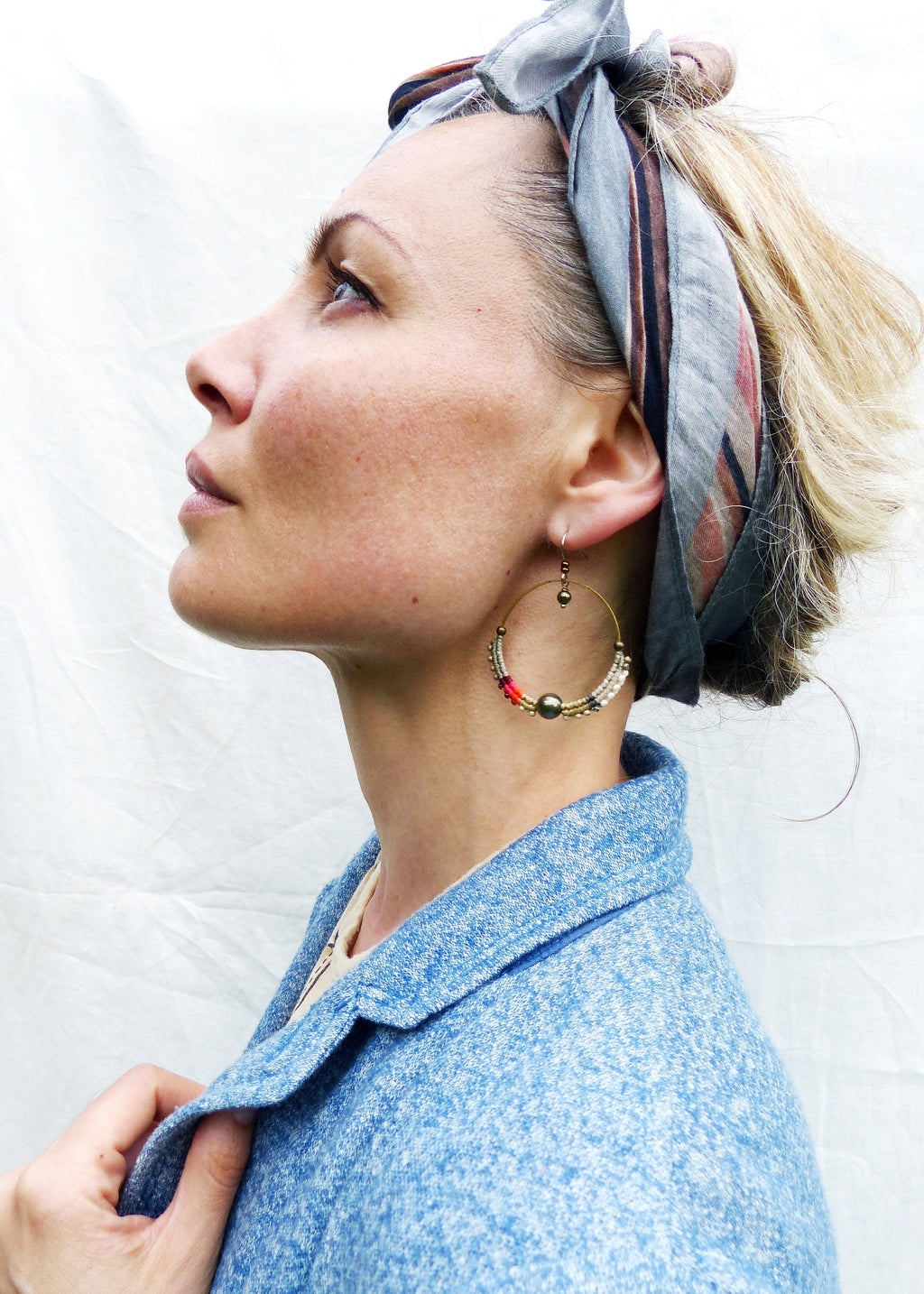 Peut inclure: Boucles d'oreilles créoles dorées avec des perles colorées et un accent de perle foncée. La personne porte une veste bleue et un foulard à motifs. Les boucles d'oreilles sont un accessoire unique.