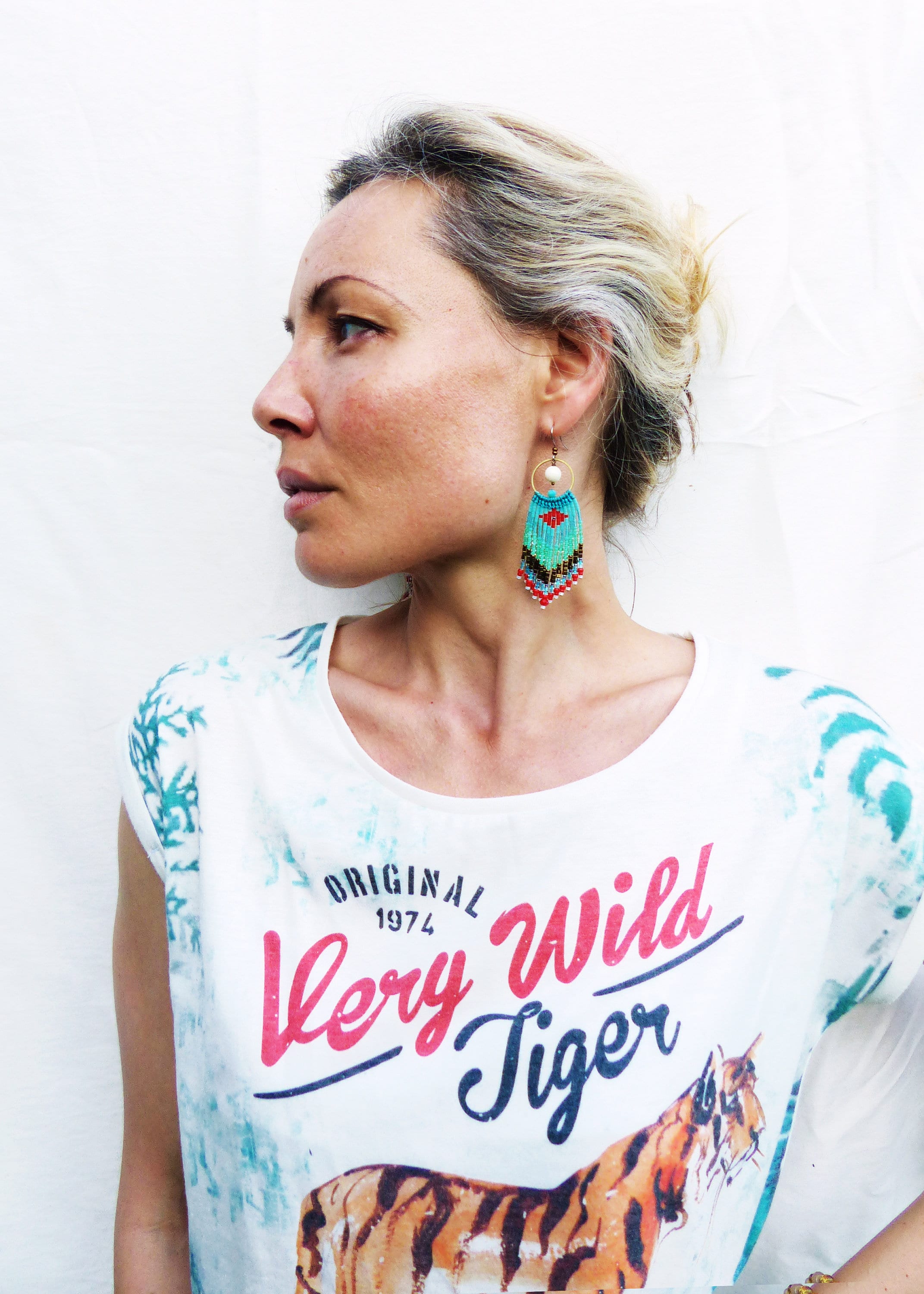 Peut inclure: Une femme portant un t-shirt blanc avec un motif de tigre et le texte "Original 1974 Very Wild Tiger". Elle porte des boucles d'oreilles en perles turquoise et rouges.