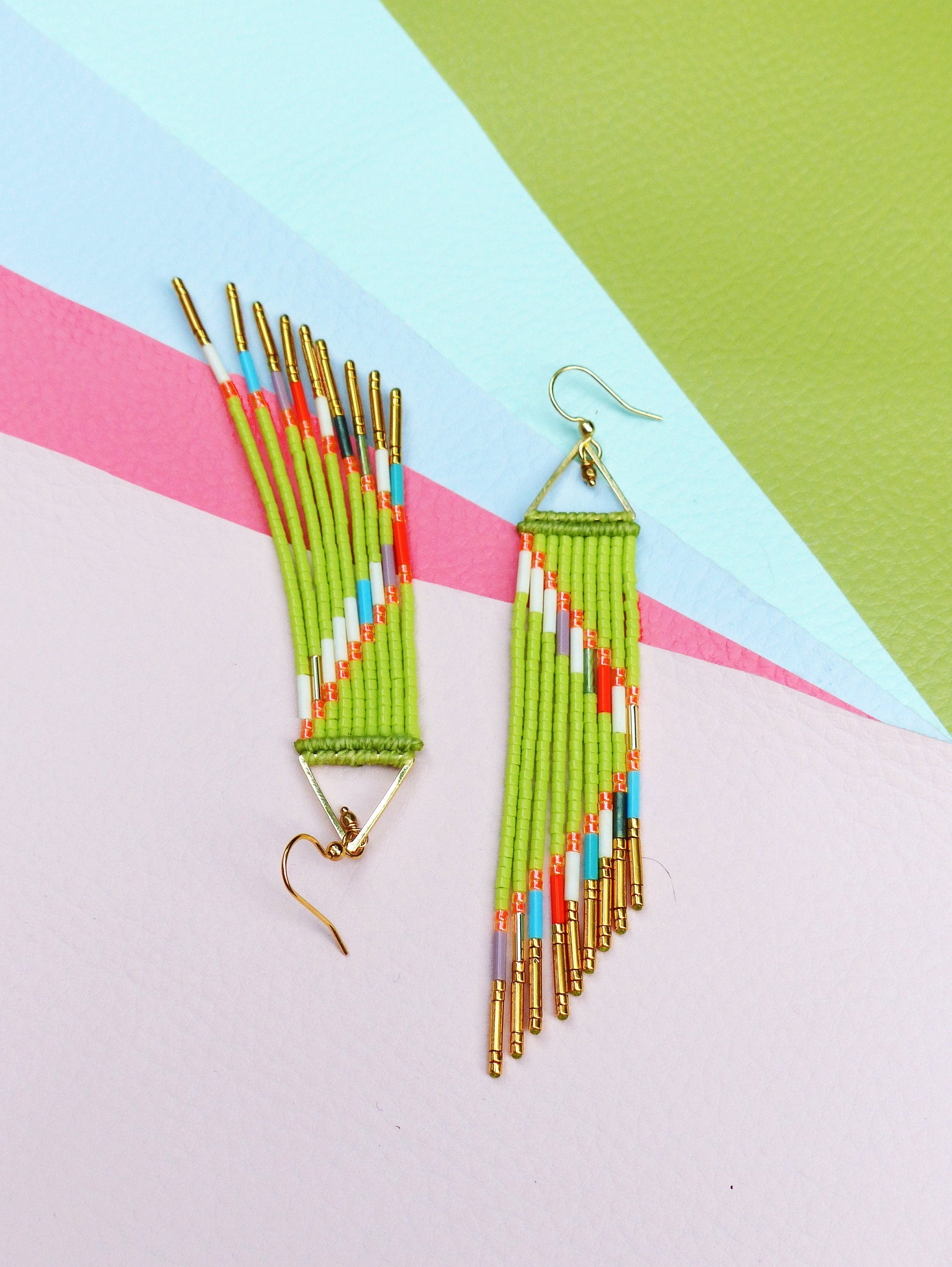 Peut inclure: Une paire de boucles d'oreilles en perles vertes avec des accents dorés et des perles colorées. Les boucles d'oreilles sont suspendues à une surface rose pâle.