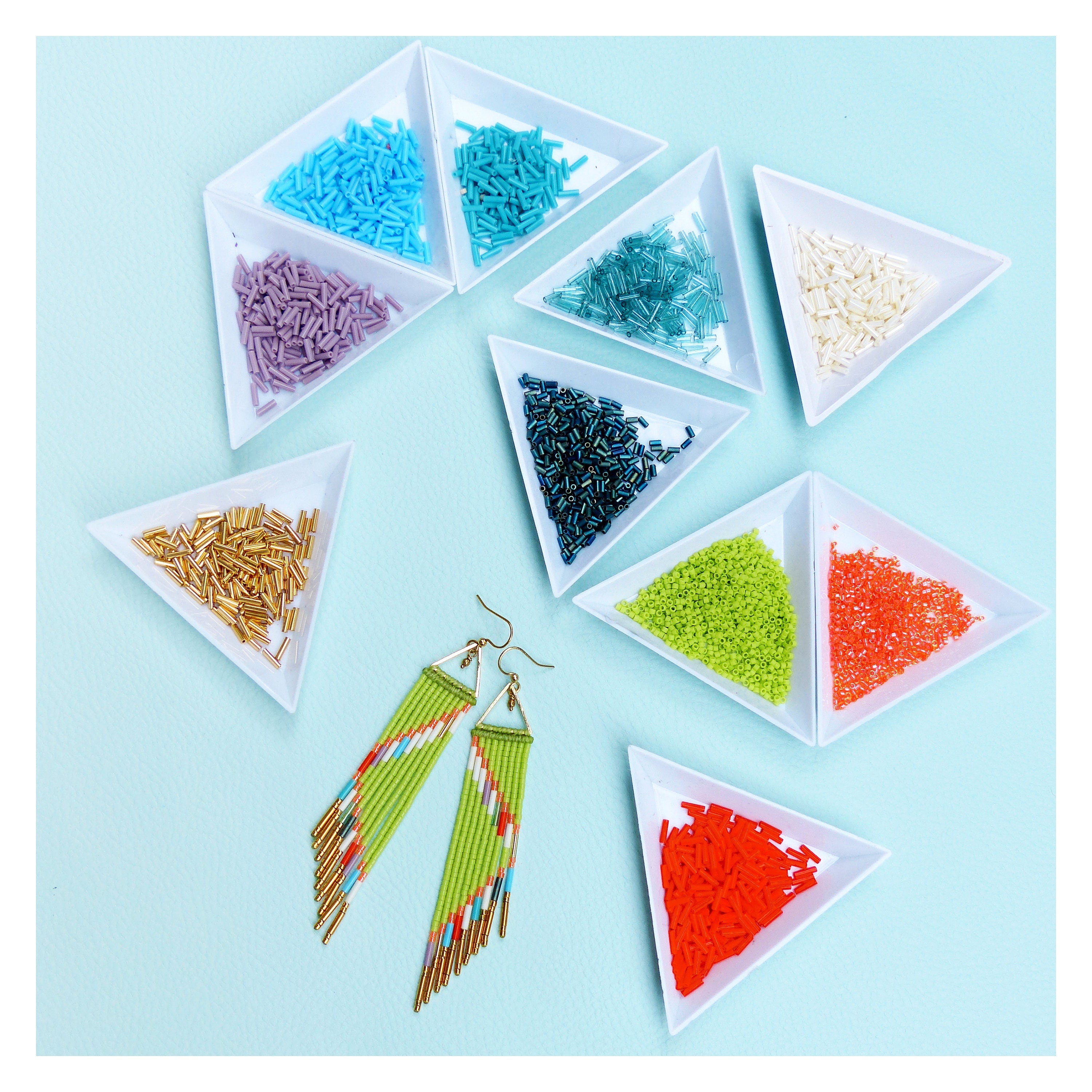 Peut inclure: Une paire de longues boucles d'oreilles en perles vertes et orange avec des accents dorés. Les boucles d'oreilles sont présentées sur une surface blanche avec de petits contenants de perles colorées.