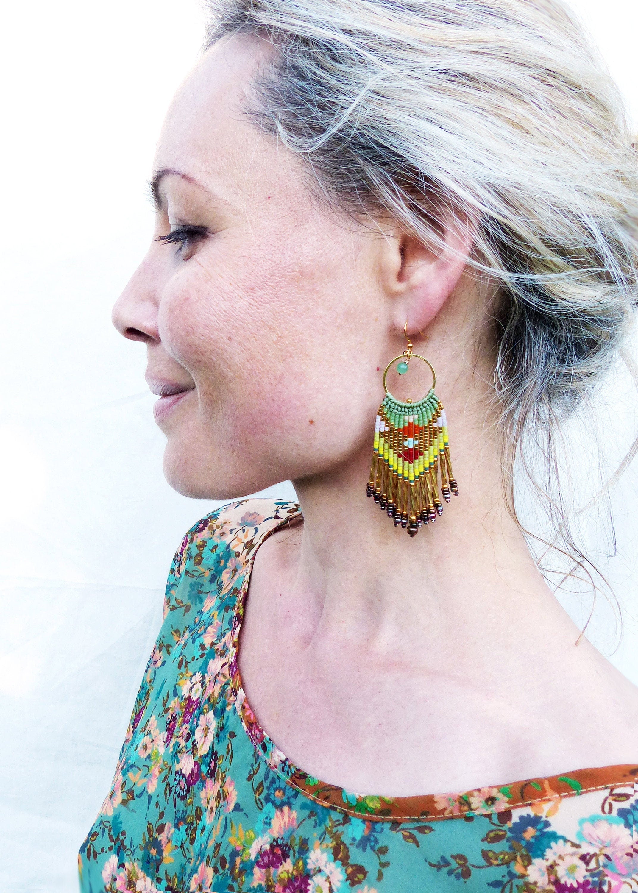 Peut inclure: Une paire de longues boucles d'oreilles en perles avec un motif géométrique dans des tons de vert, jaune, orange et marron. Les boucles d'oreilles sont suspendues à un anneau en or et présentent un petit accent de pierre verte.