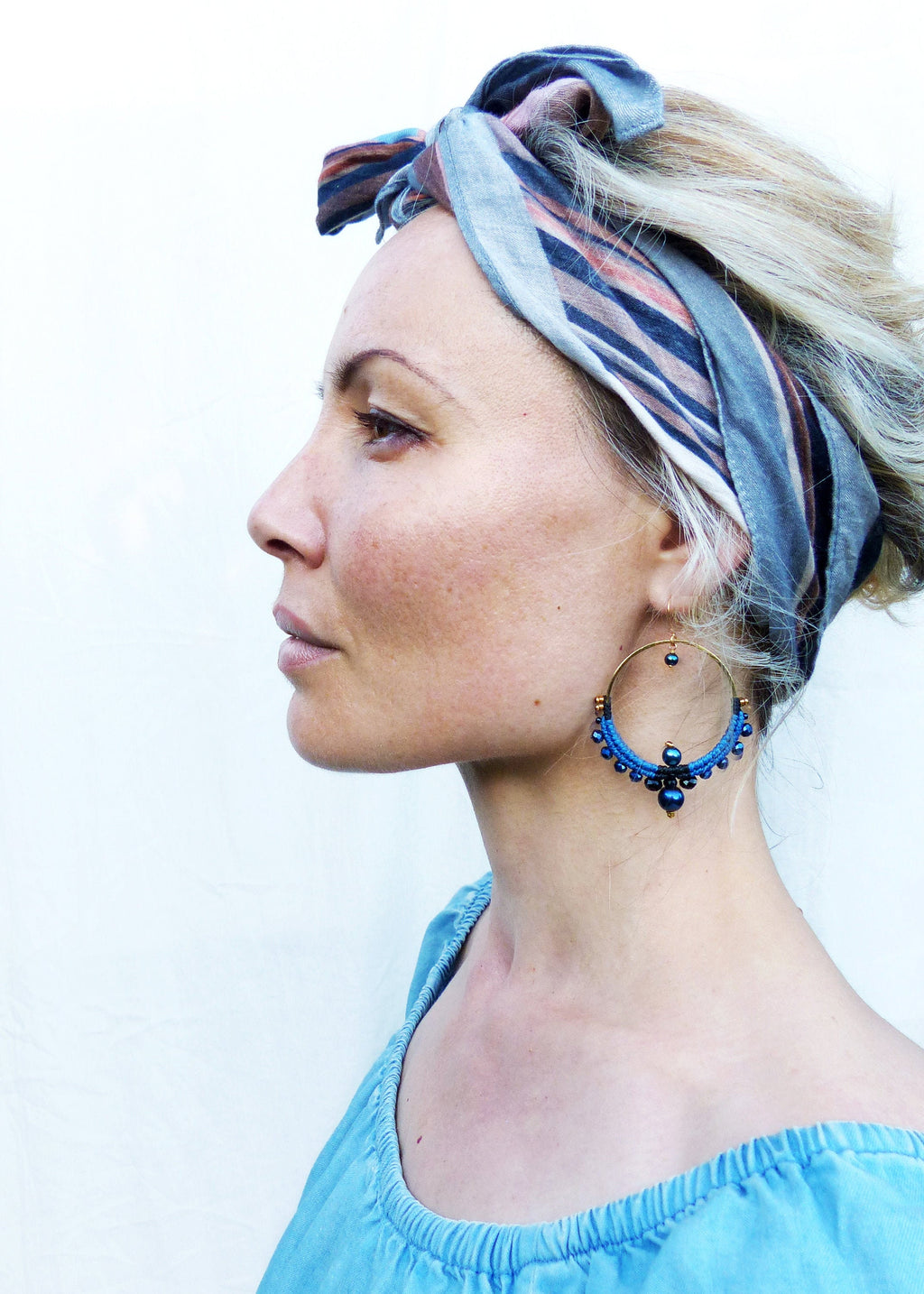 Peut inclure: Une femme aux cheveux blonds porte un bandeau rayé bleu et marron et de grandes boucles d'oreilles créoles bleues avec des perles bleu foncé. Les boucles d'oreilles ont un anneau en métal doré.