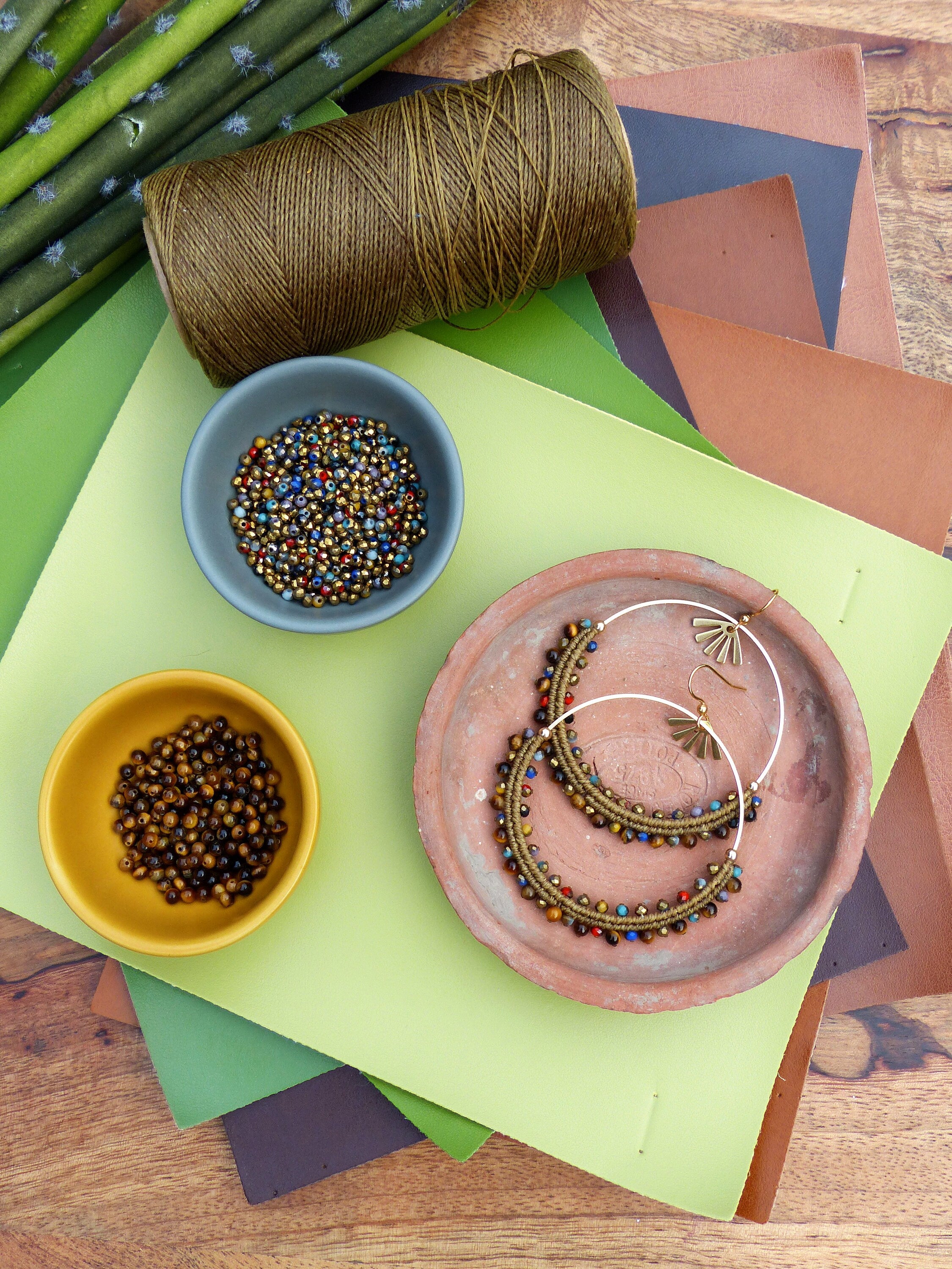 Peut inclure: Une paire de boucles d'oreilles créoles brunes et dorées avec des perles colorées, une bobine de fil brun et deux bols de perles sur un fond vert et brun.