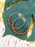 Peut inclure: Une paire de boucles d'oreilles créoles dorées avec des perles brunes et multicolores. Les boucles d'oreilles sont sur une feuille verte.