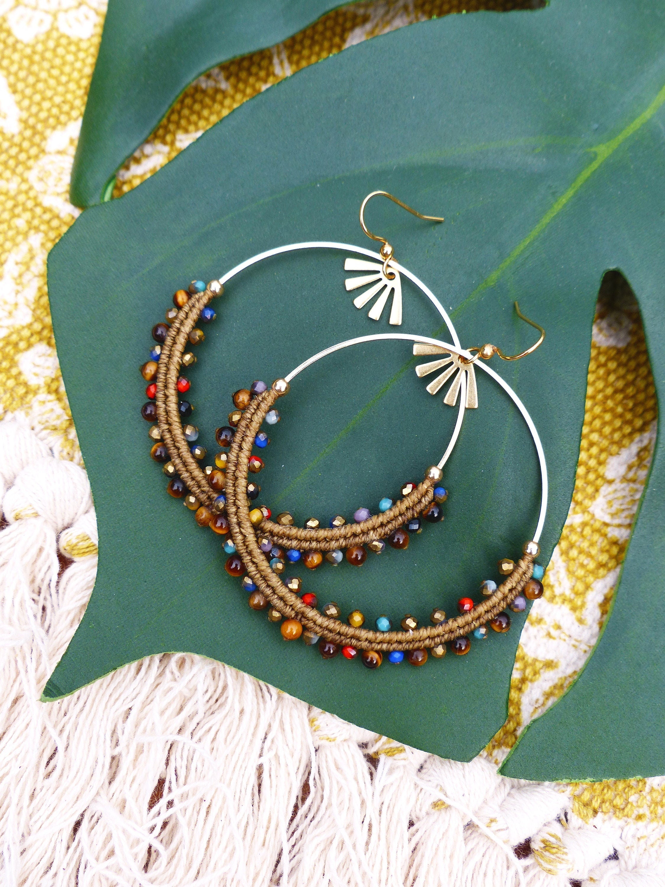 Peut inclure: Une paire de boucles d'oreilles créoles dorées avec des perles brunes et multicolores. Les boucles d'oreilles sont sur une feuille verte.