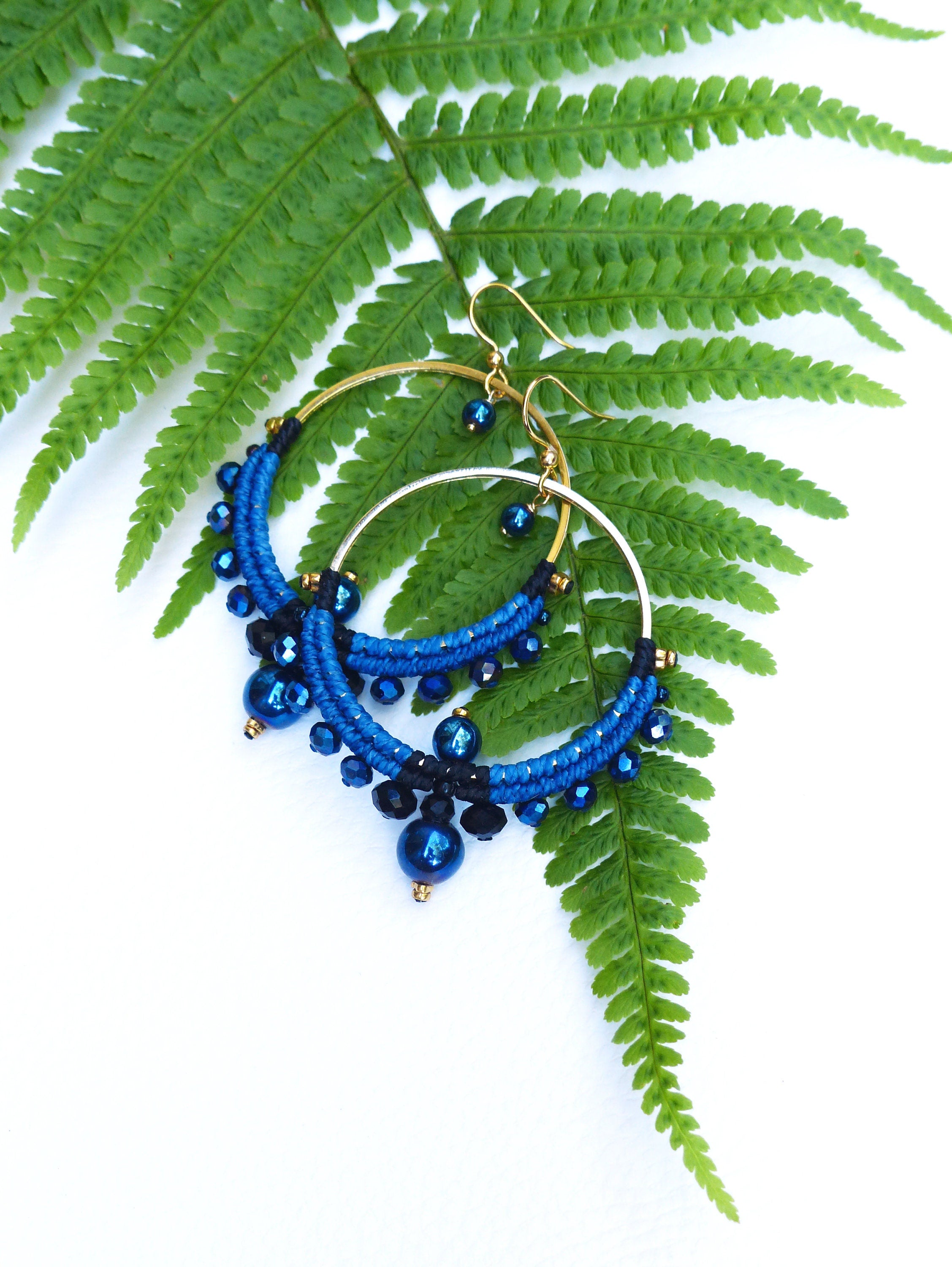 Peut inclure: Une paire de boucles d'oreilles créoles en perles bleues et noires avec des cercles dorés. Les boucles d'oreilles sont suspendues à une feuille de fougère verte.