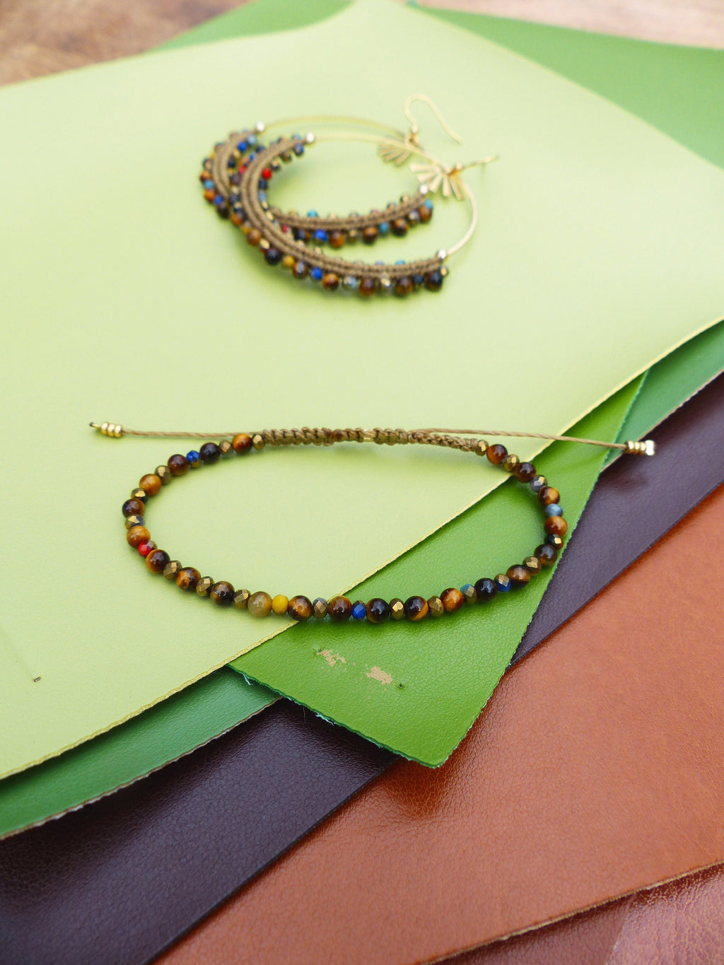 Peut inclure: Un bracelet en perles brunes et dorées avec un fermoir doré. Le bracelet est fait de perles en œil de tigre et se trouve sur un fond vert et brun.
