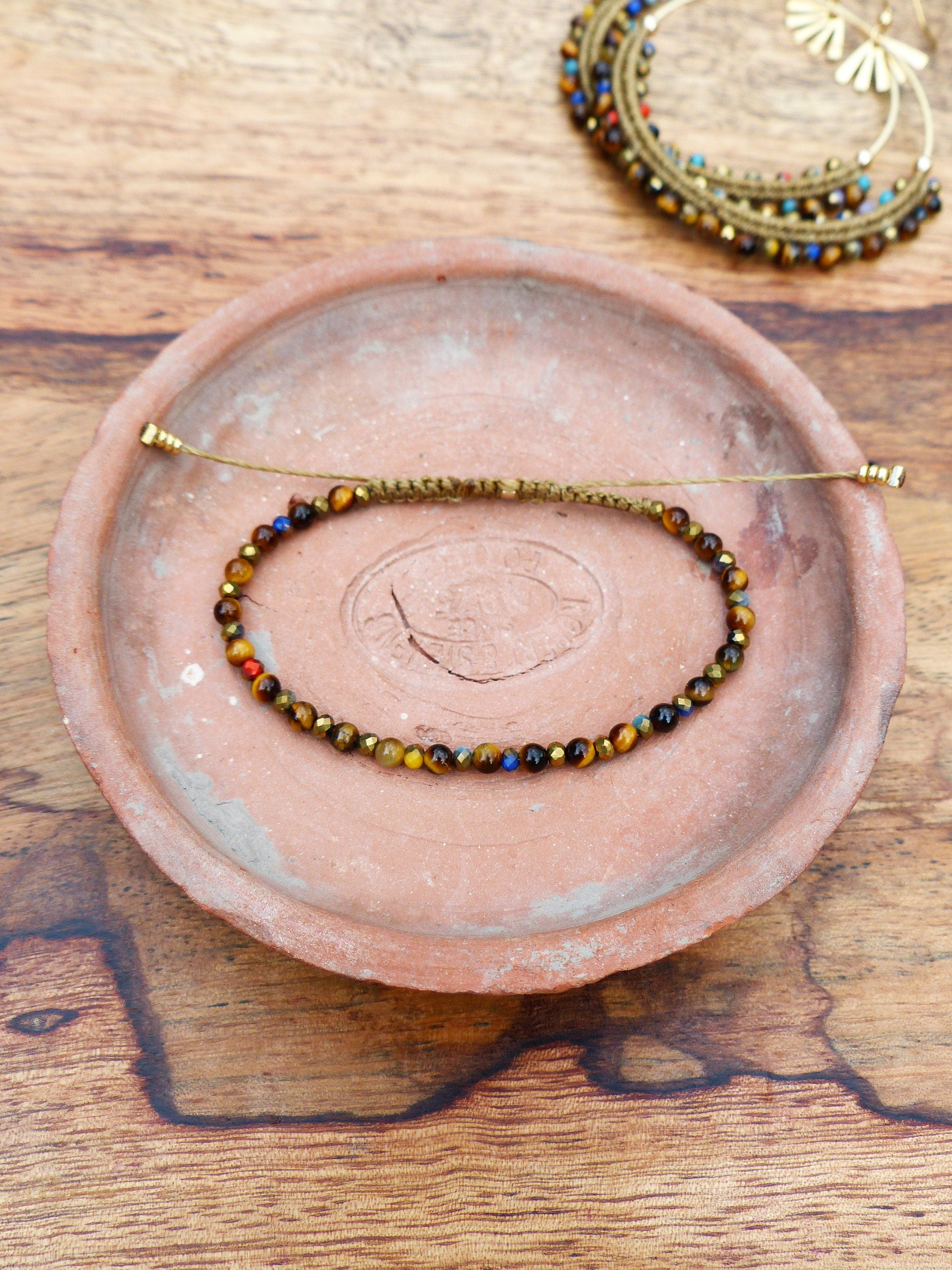 Peut inclure: Un bracelet en perles brunes, dorées et bleues avec un cordon brun et des embouts dorés. Le bracelet est sur une assiette en terre cuite.