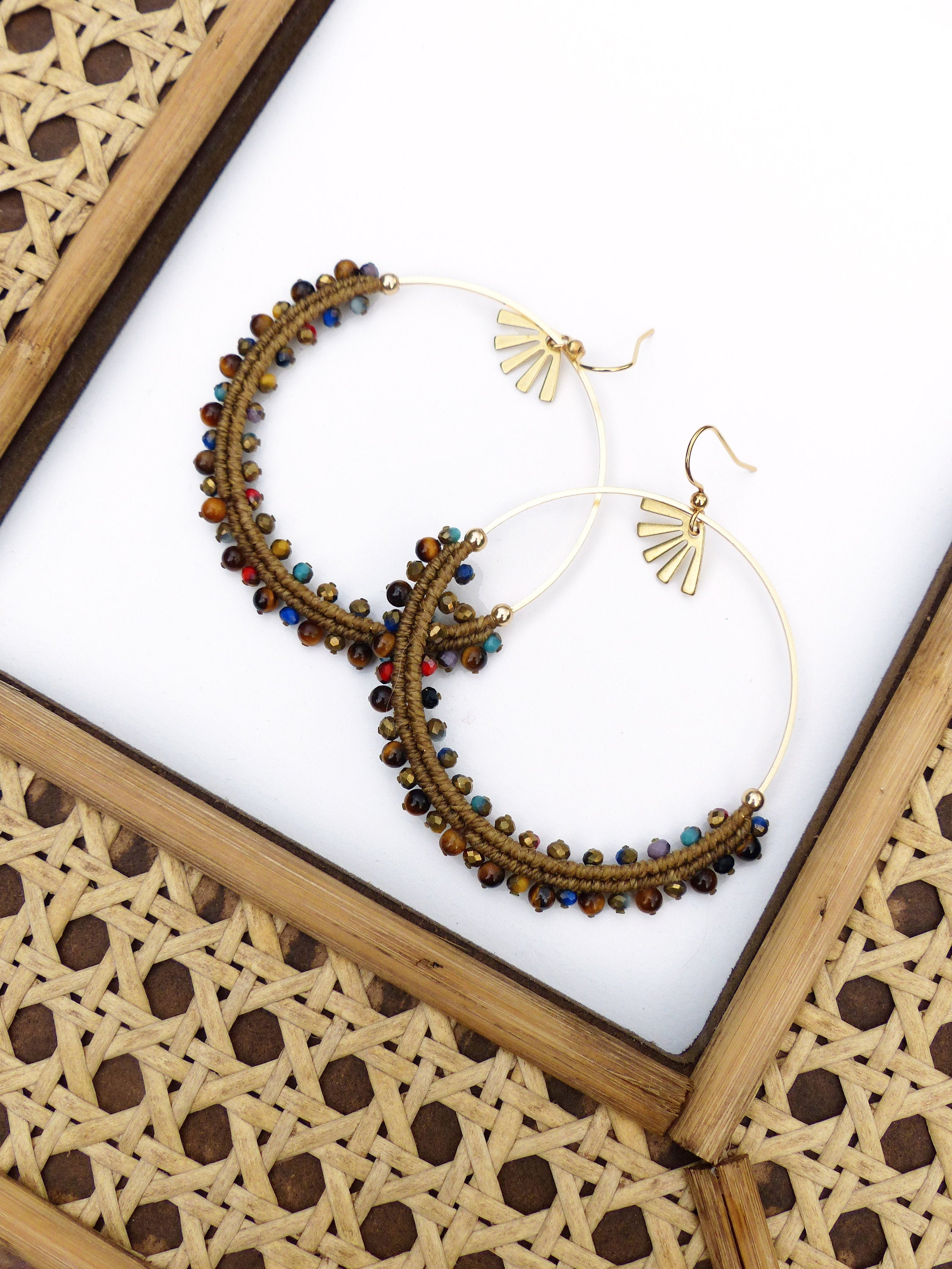 Peut inclure: Une paire de boucles d'oreilles créoles dorées avec des perles brunes et multicolores. Les boucles d'oreilles ont un motif en forme d'éventail en haut.