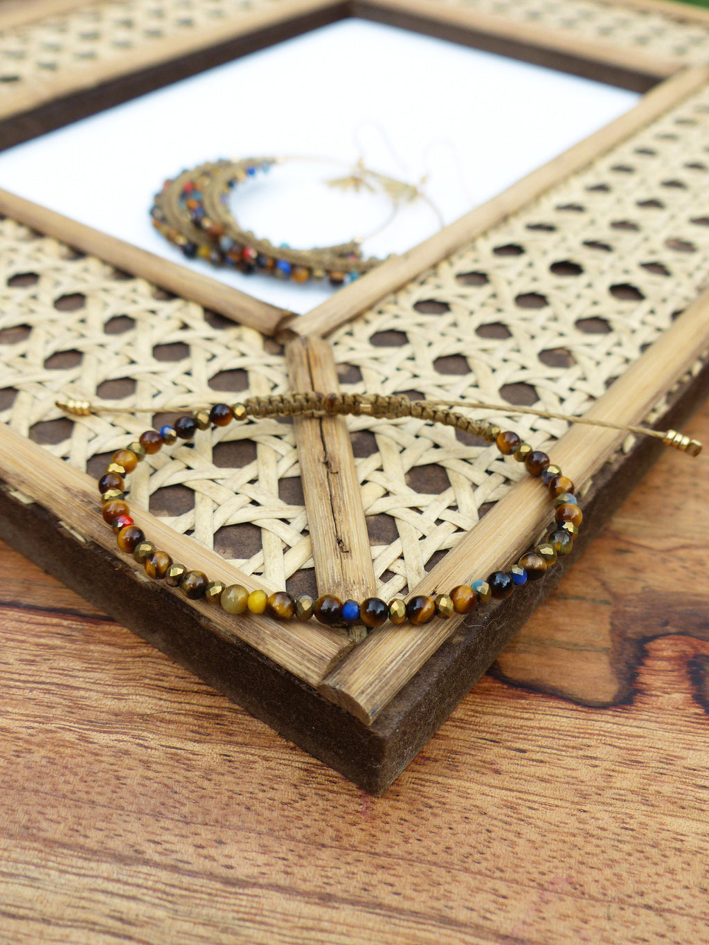 Peut inclure: Un bracelet en perles brunes et dorées avec une fermeture en cordon tissé. Le bracelet est composé de perles en œil de tigre avec des accents bleus et rouges. Le bracelet est présenté sur un cadre en bambou tissé.