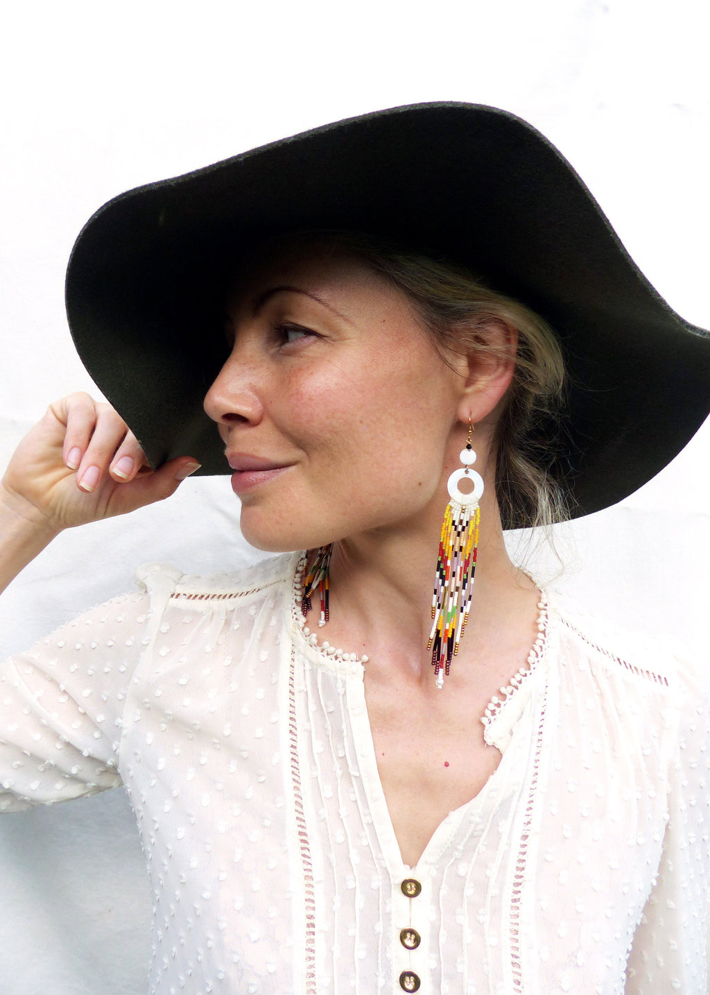 Peut inclure: Une femme portant une blouse blanche avec un chapeau noir à larges bords et de longues boucles d'oreilles en perles blanches et colorées.
