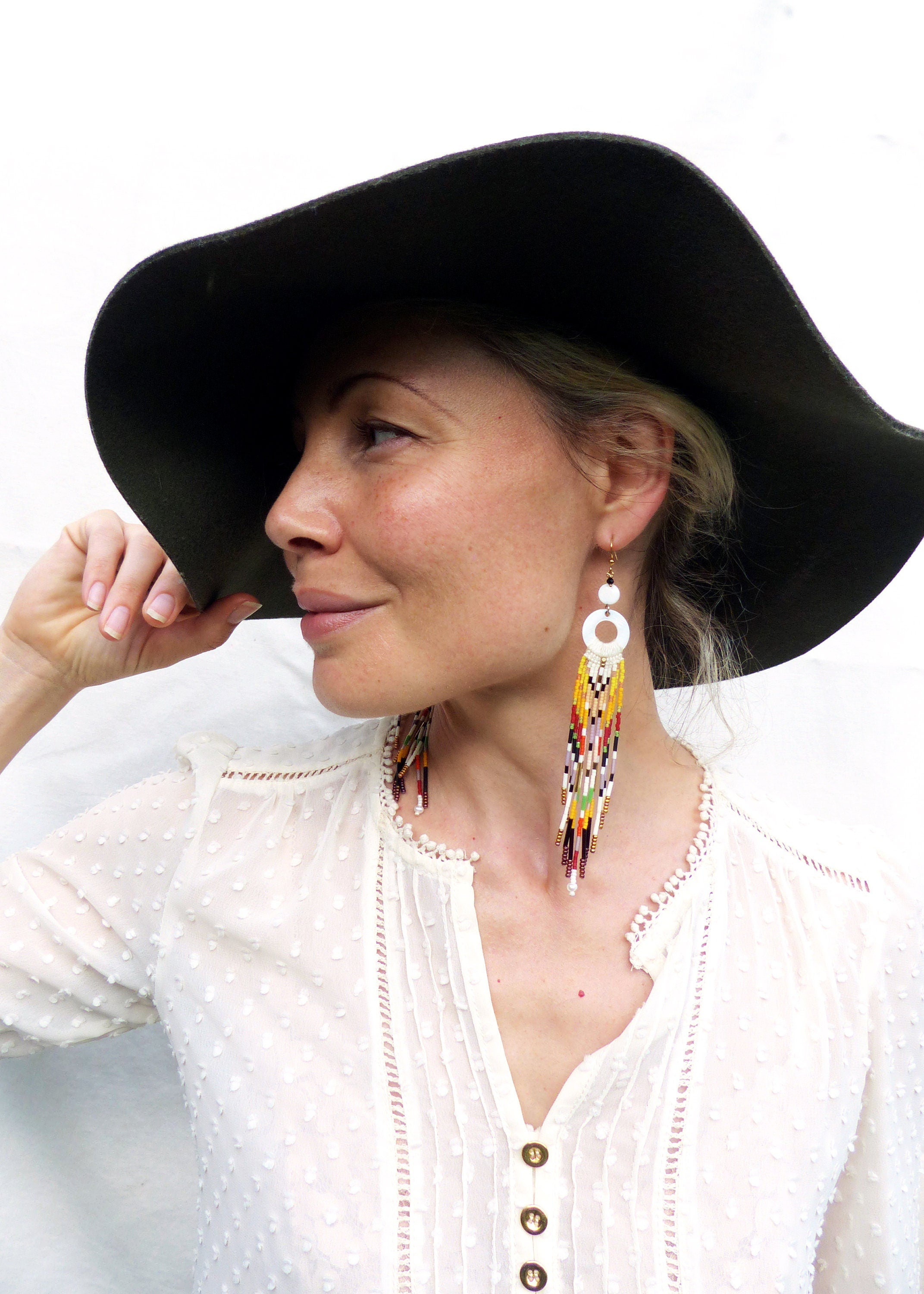 Peut inclure: Une femme portant une blouse blanche avec un chapeau noir à larges bords et de longues boucles d'oreilles en perles blanches et colorées.