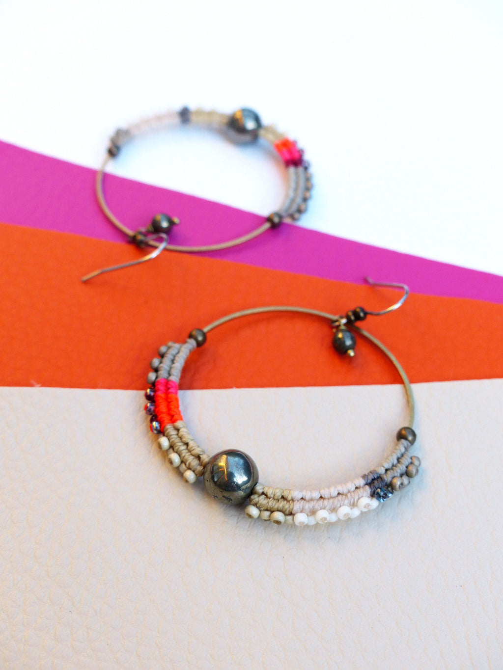 Peut inclure: Deux boucles d'oreilles créoles avec une monture en métal et des détails perlés. Les boucles d'oreilles comportent une grosse perle gris foncé au centre et sont ornées de perles grises, roses et blanches. Les boucles d'oreilles sont posées sur un fond coloré.