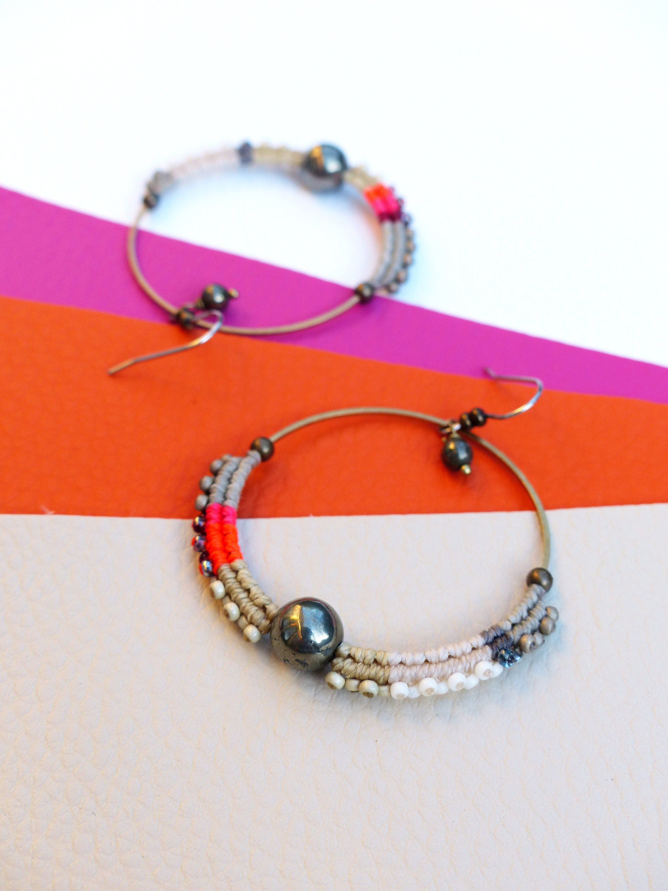 Peut inclure: Deux boucles d'oreilles créoles avec une monture en métal et des détails perlés. Les boucles d'oreilles comportent une grosse perle gris foncé au centre et sont ornées de perles grises, roses et blanches. Les boucles d'oreilles sont posées sur un fond coloré.