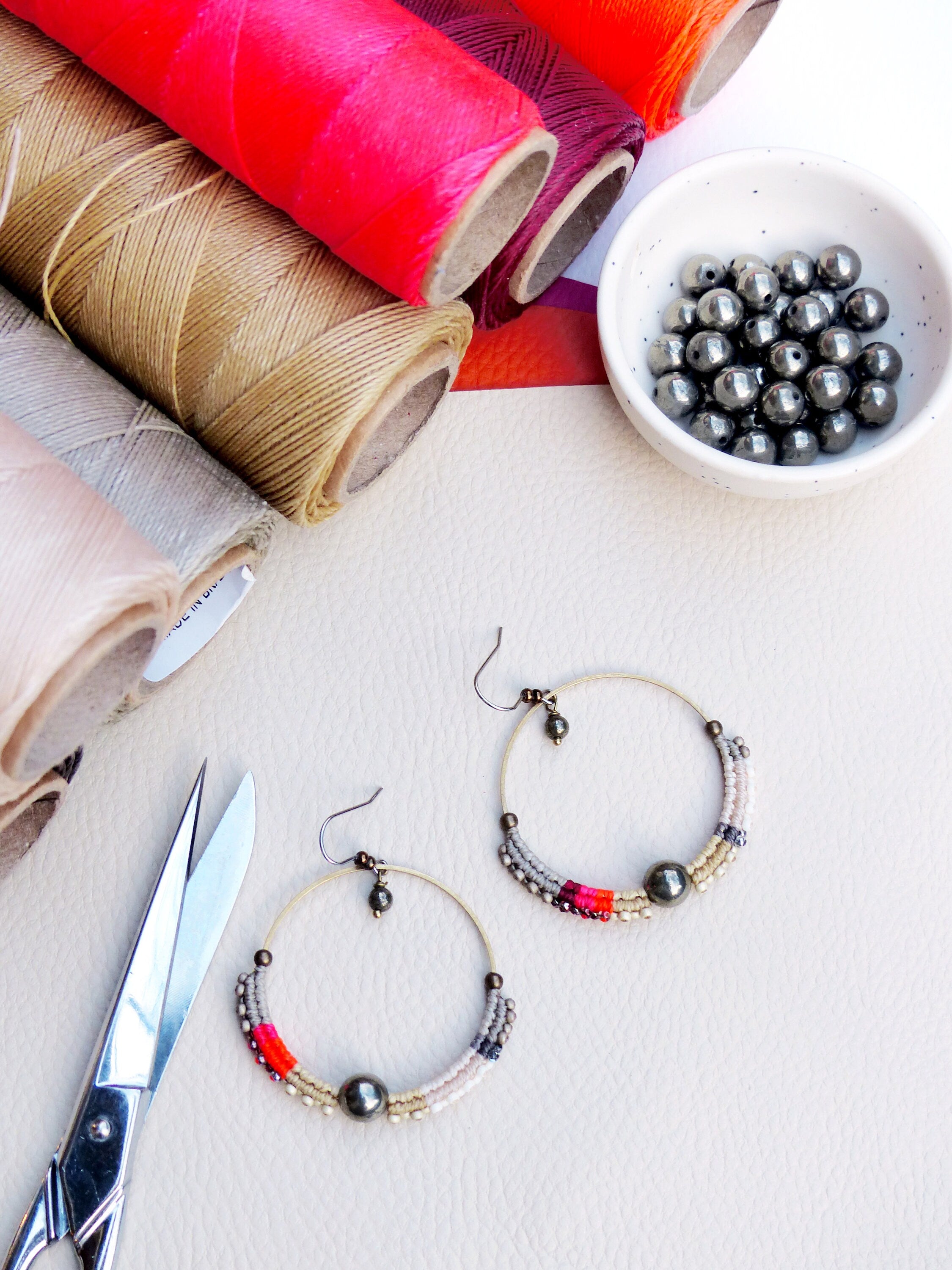 Peut inclure: Boucles d'oreilles créoles faites à la main avec un design perlé, aux tons de rouge, rose, gris et crème. Les boucles d'oreilles sont présentées avec des bobines de fil coloré, un petit bol de perles et des ciseaux, suggérant un thème artisanal. Diamètre: environ 5 cm.