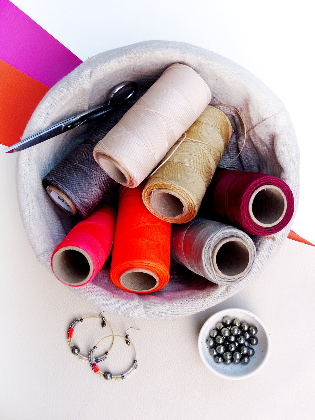 Peut inclure: Un assortiment de fournitures de couture comprenant des bobines de fil de différentes couleurs, une paire de ciseaux et un petit bol de perles. Les couleurs des fils varient du beige au rouge et au gris. Des boucles d'oreilles créoles avec des perles colorées sont également présentes.