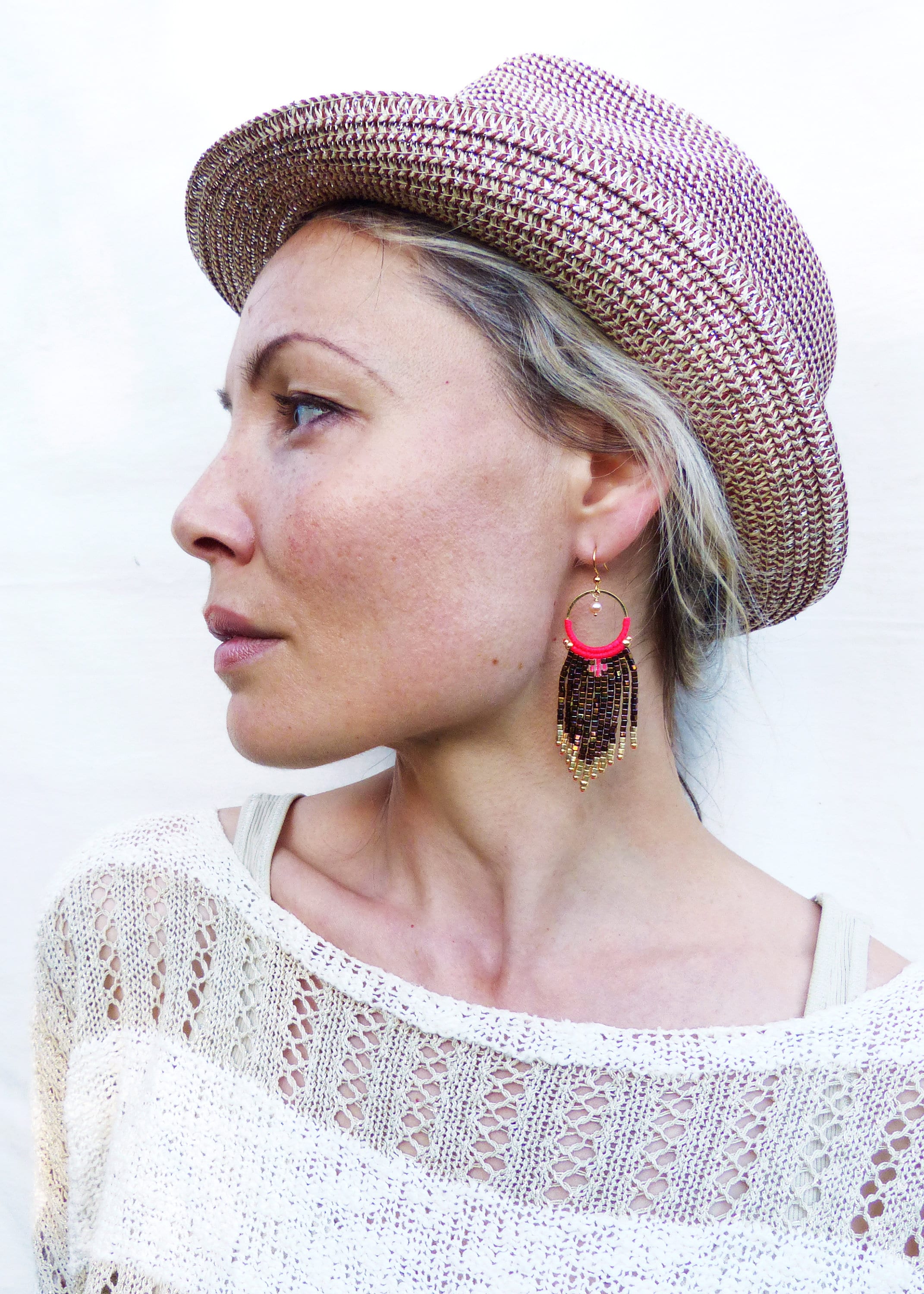 Peut inclure: Une femme portant un chapeau de paille brun et blanc avec une boucle d'oreille à franges en perles brunes et dorées. La boucle d'oreille a un accent rouge.