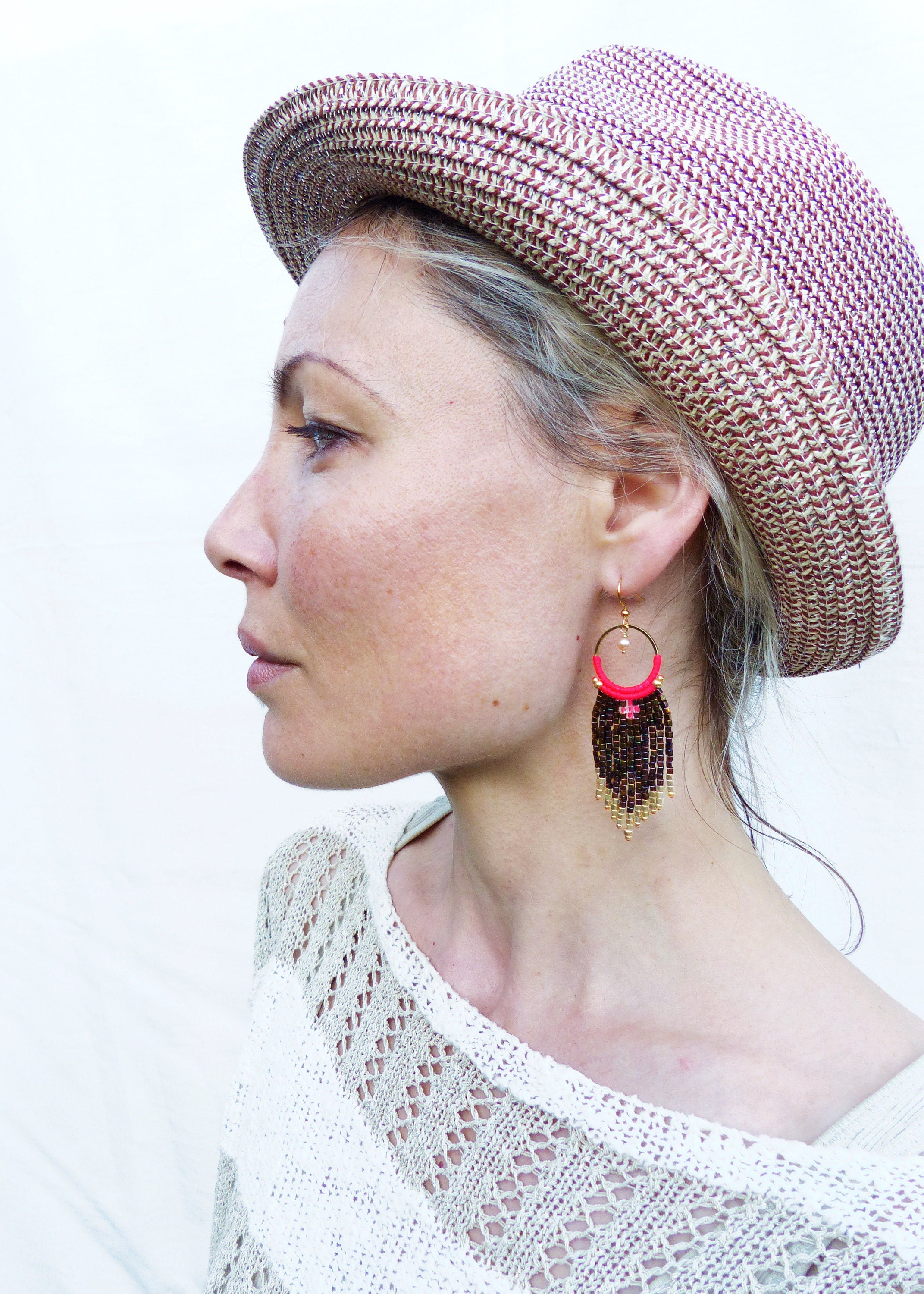 Peut inclure: Une femme portant un chapeau de paille brun et blanc à large bord et un haut en crochet blanc. Elle porte de longues boucles d'oreilles en perles avec un motif rouge et or.