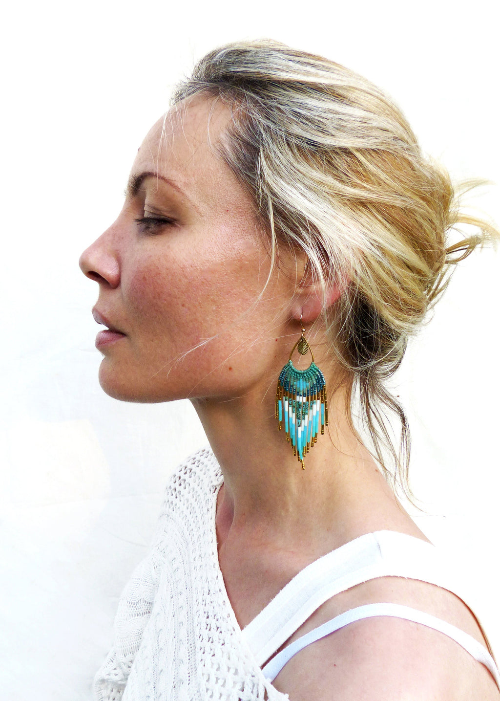 Peut inclure: Profil d'une personne portant une boucle d'oreille en perles turquoise, blanches et dorées. La boucle d'oreille a une forme de ventilateur avec un dessus en métal doré. La personne porte un haut blanc.