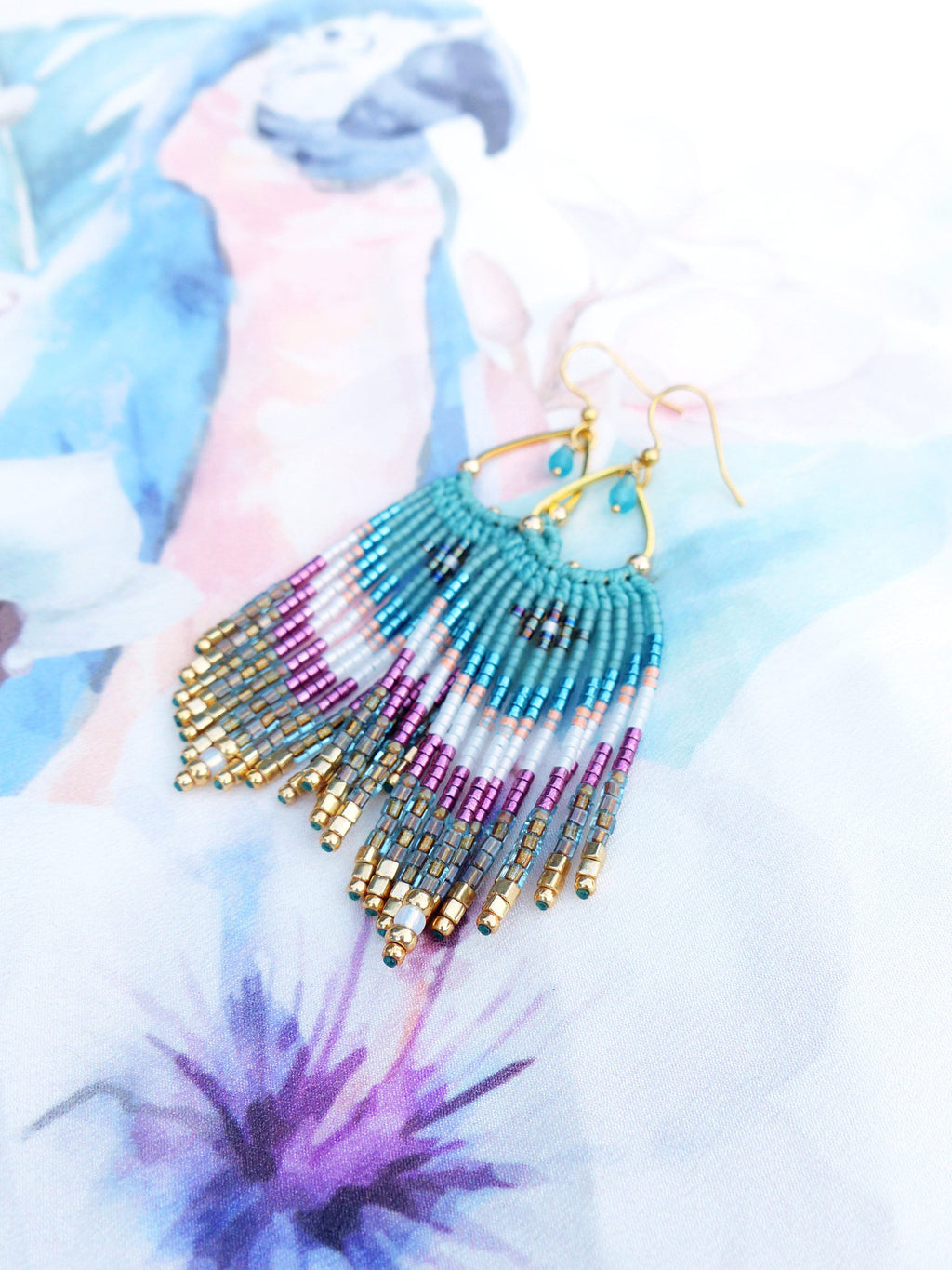 Peut inclure: Une paire de boucles d'oreilles pendantes en perles turquoise, blanches et violettes avec des cercles dorés et une perle turquoise en forme de larme en haut.
