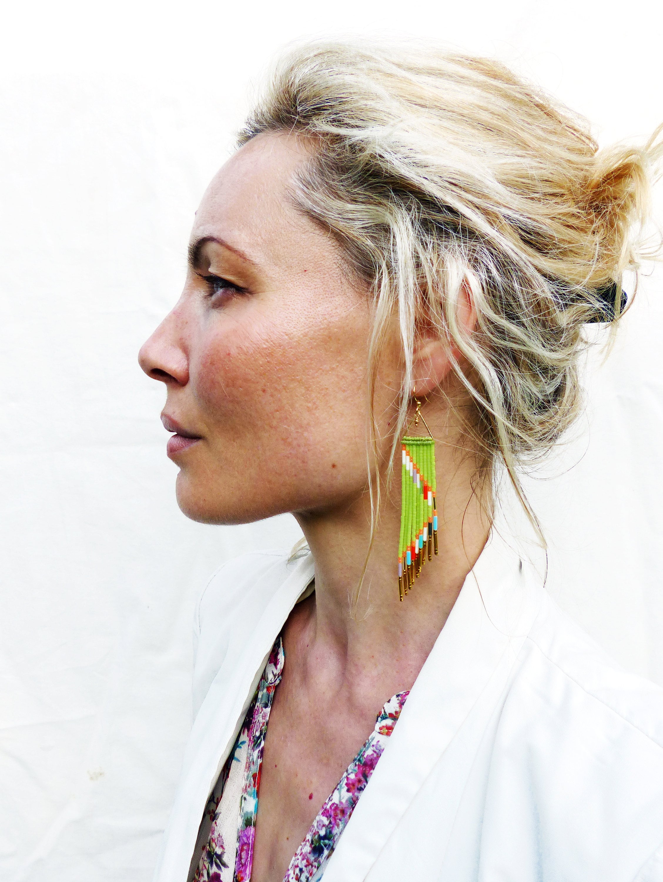 Peut inclure: Une femme aux cheveux blonds portant un blazer blanc et une blouse à fleurs. Elle porte de longues boucles d'oreilles en perles vertes et dorées.