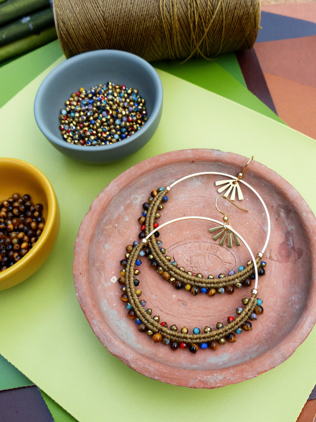 Peut inclure: Une paire de grandes boucles d'oreilles créoles avec un cordon brun enroulé autour du cercle et rehaussé de perles colorées. Les boucles d'oreilles sont présentées sur une assiette en terre cuite.