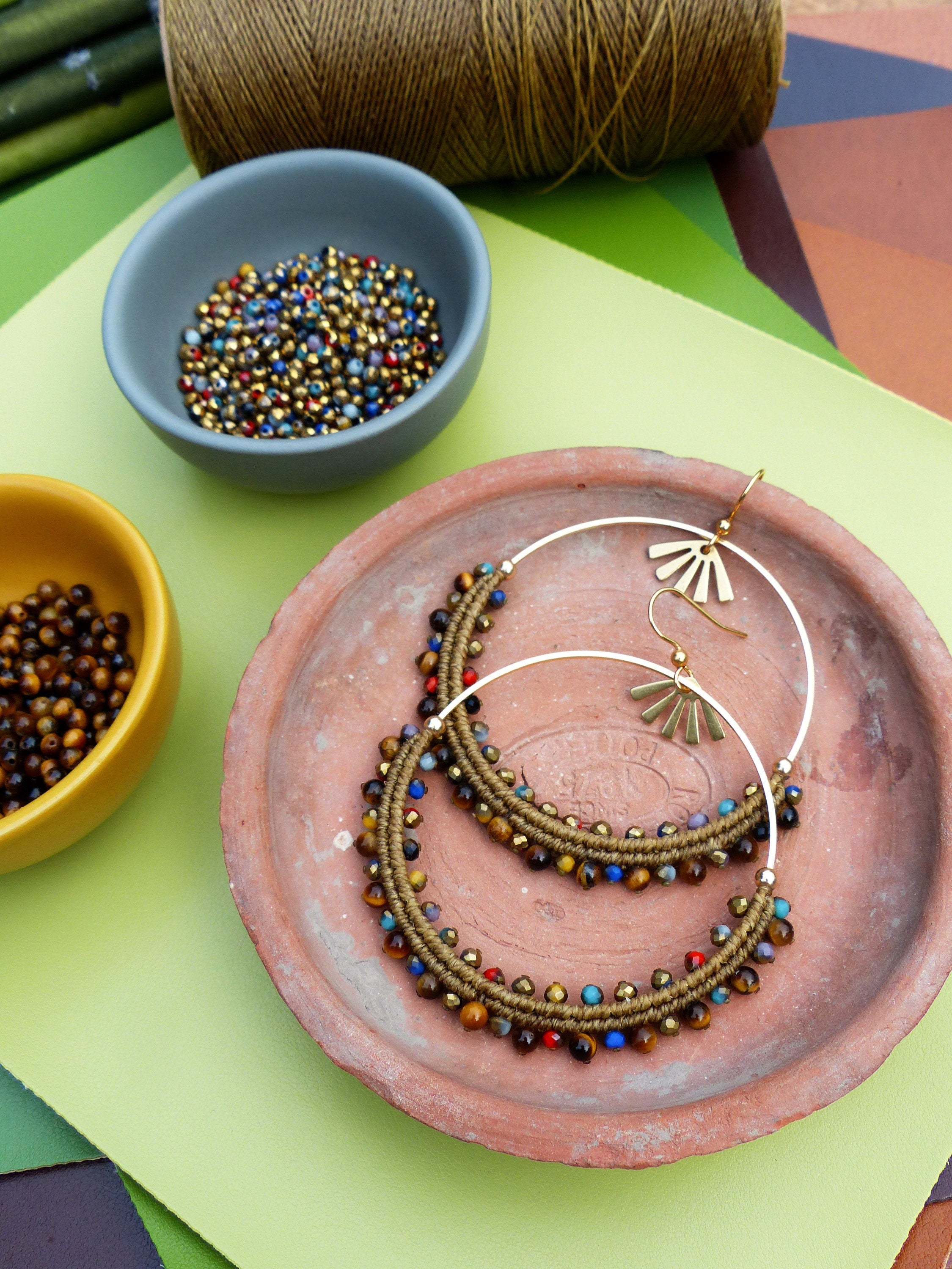 Peut inclure: Une paire de grandes boucles d'oreilles créoles avec un cordon brun enroulé autour du cercle et rehaussé de perles colorées. Les boucles d'oreilles sont présentées sur une assiette en terre cuite.