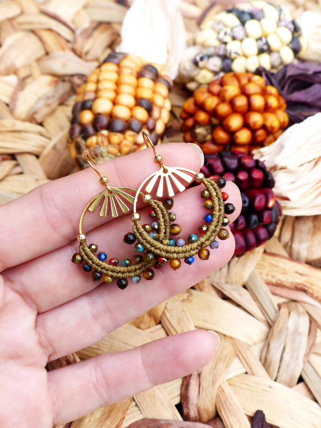 Peut inclure: Une paire de boucles d'oreilles créoles dorées avec une frange de perles. Les perles sont brunes, vertes, bleues et rouges. Les boucles d'oreilles sont suspendues à une main.