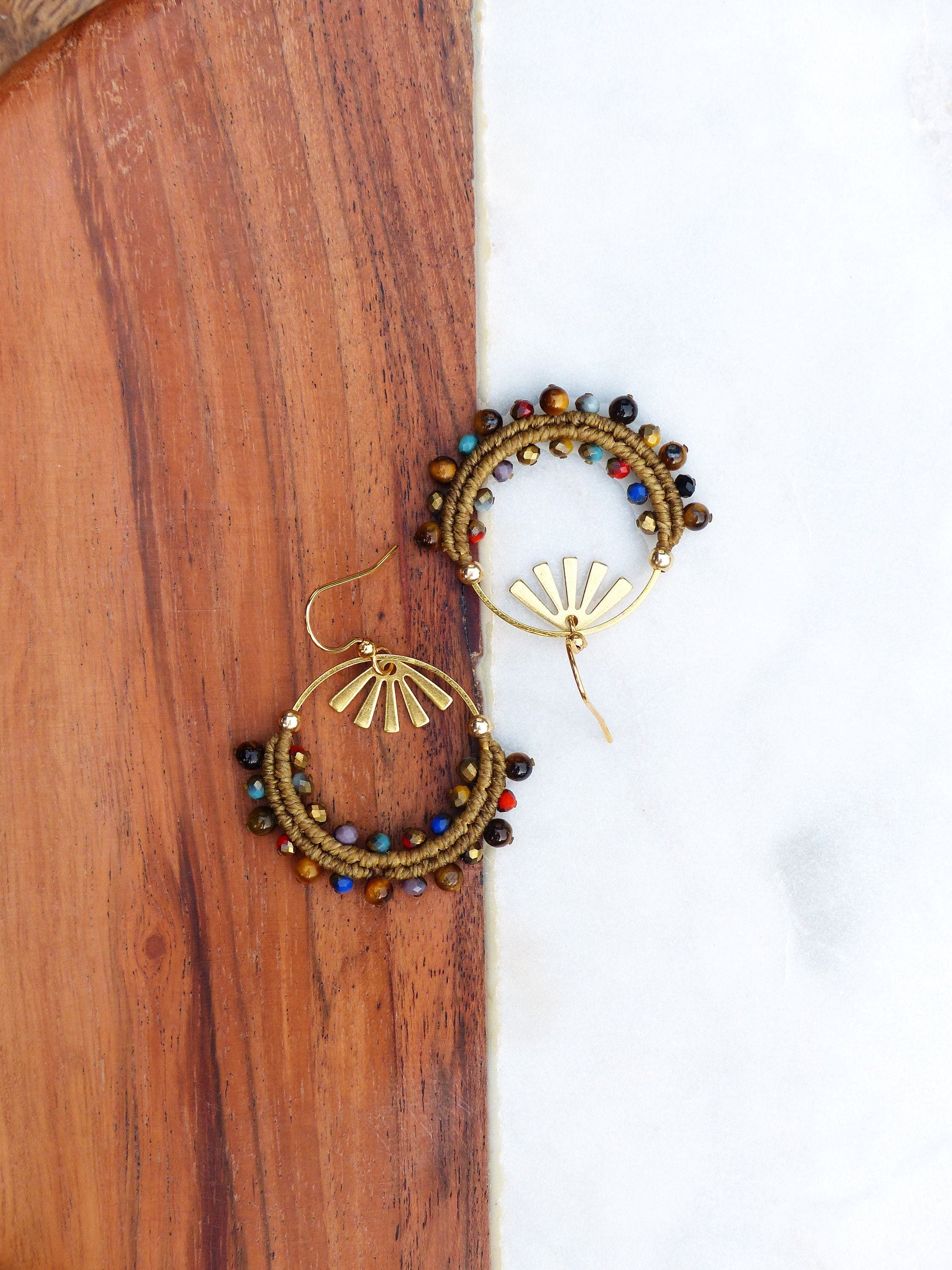 Peut inclure: Une paire de boucles d'oreilles créoles dorées avec un motif soleil et des perles multicolores. Les perles sont brunes, rouges, bleues, vertes et jaunes.