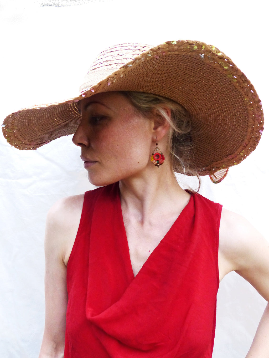 Peut inclure: Une femme portant un haut rouge sans manches et un chapeau de paille à larges bords avec une bordure rose et dorée. Elle porte une paire de boucles d'oreilles pendantes avec des accents rouges, orange et jaunes.