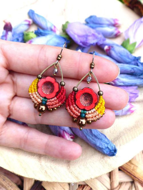 Peut inclure: Une paire de boucles d'oreilles colorées et faites à la main avec un motif de fleur rouge. Les boucles d'oreilles sont en macramé et en perles, et ont une finition dorée.