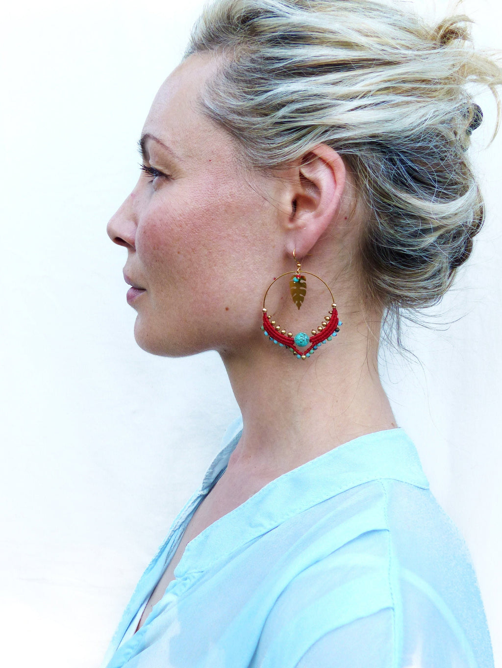 Peut inclure: Une femme portant une chemise bleu clair avec une paire de boucles d'oreilles créoles rouges et turquoise avec un pendentif en feuille d'or.