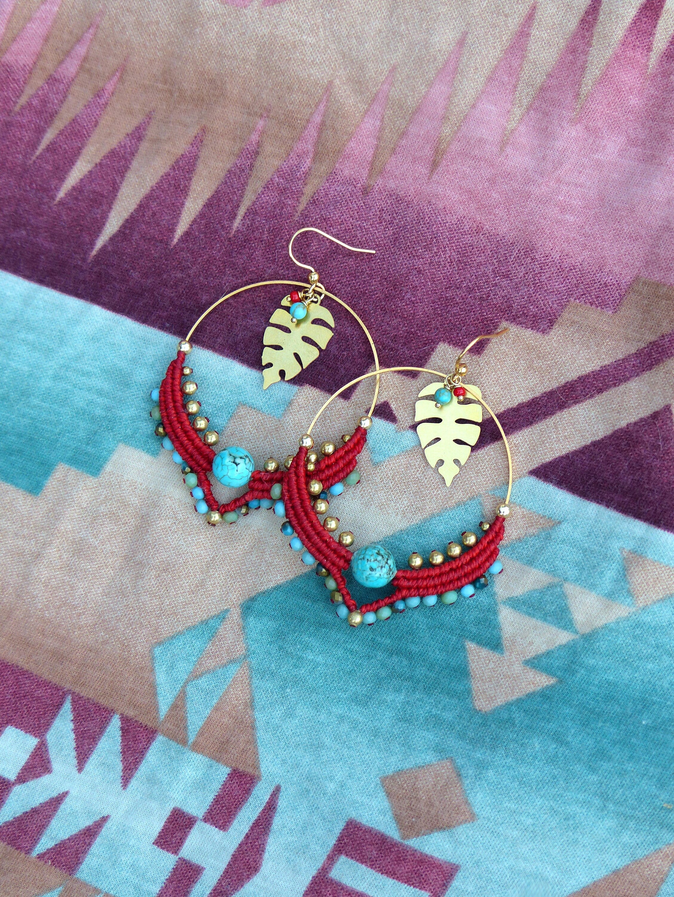 Peut inclure: Une paire de boucles d'oreilles créoles dorées avec des perles rouges et turquoise et un pendentif en forme de feuille dorée. Les boucles d'oreilles sont sur un fond de tissu à motifs.