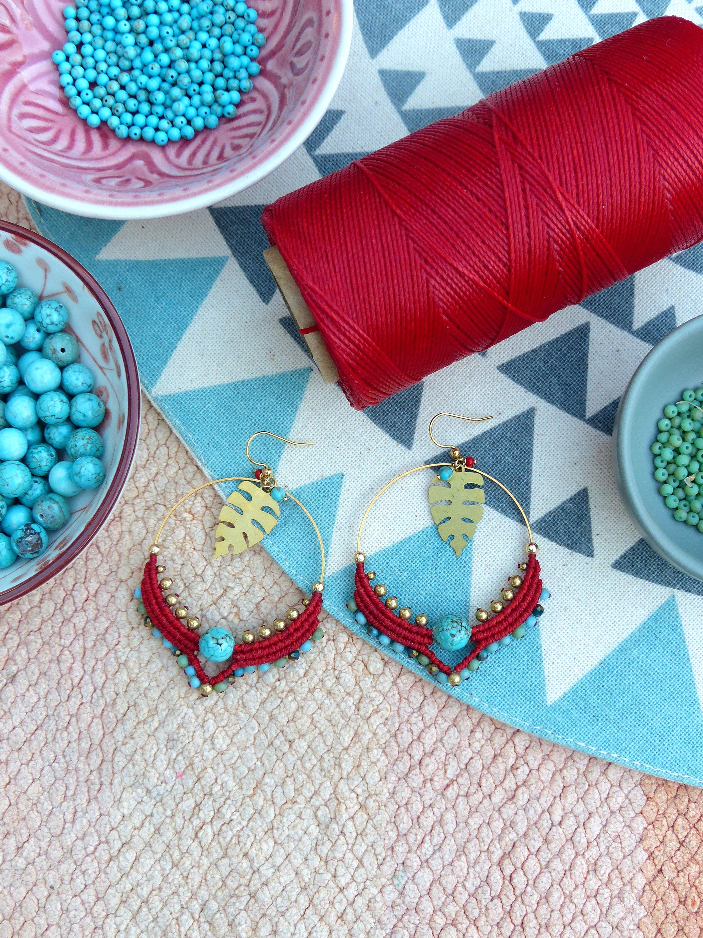 Peut inclure: Une paire de boucles d'oreilles créoles rouges et dorées avec des perles turquoise et des breloques en forme de feuilles. Les boucles d'oreilles sont sur une surface à motifs blancs et bleus avec une bobine de fil rouge et des bols de perles turquoise.