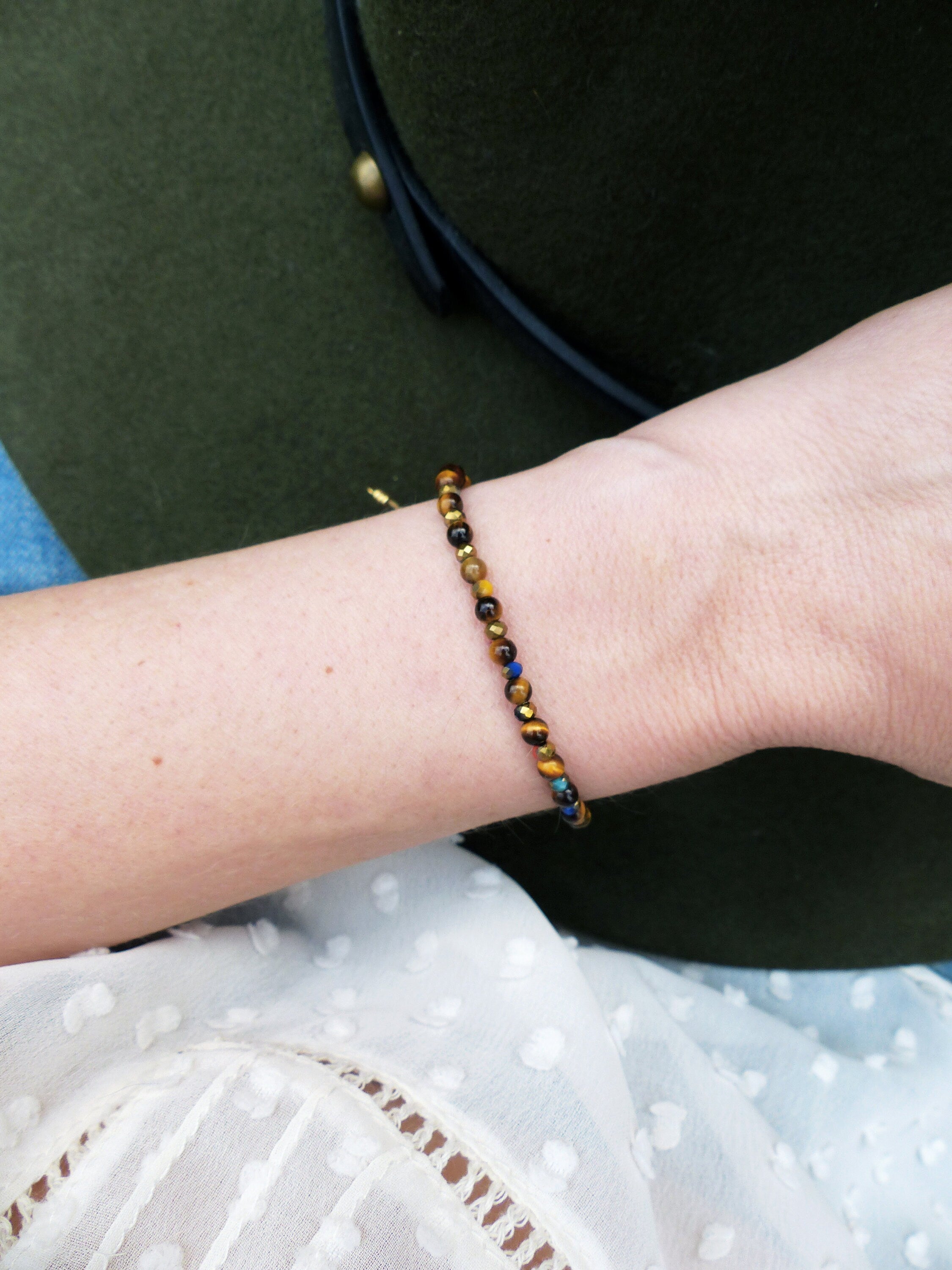 Peut inclure: Un bracelet en or avec des perles brunes, noires et bleues. Le bracelet est porté au poignet.