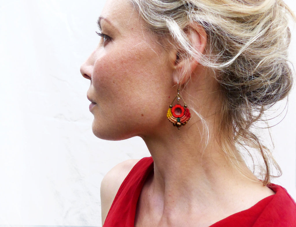 Peut inclure: Une paire de boucles d'oreilles en perles rouges, orange et jaunes avec un élément central rond rouge. Les boucles d'oreilles pendent de l'oreille d'une personne aux cheveux blonds.