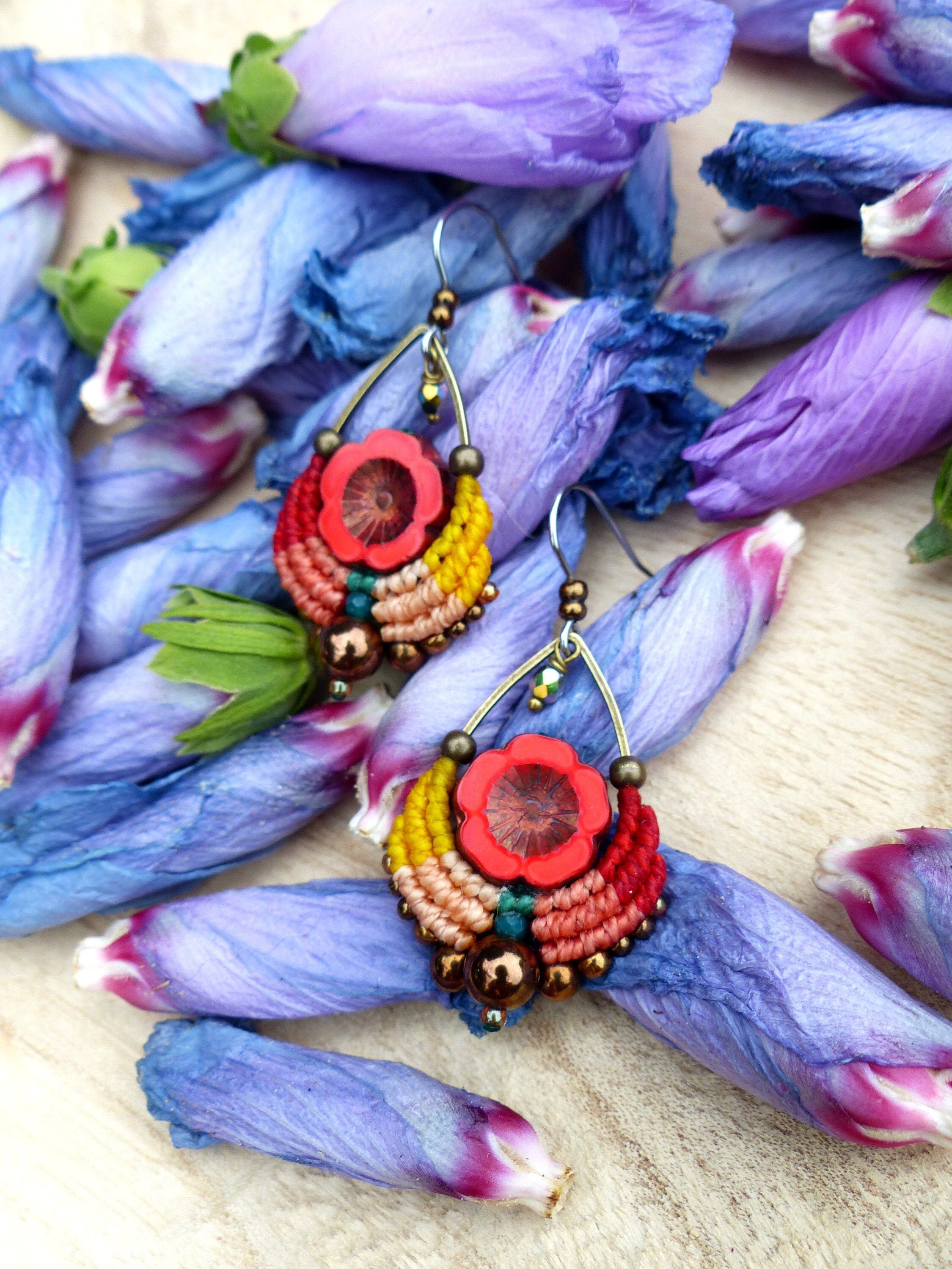 Peut inclure: Une paire de boucles d'oreilles colorées avec un design en macramé rouge, orange, jaune et vert. Les boucles d'oreilles présentent une perle rouge en forme de fleur au centre et sont rehaussées de perles bronze.