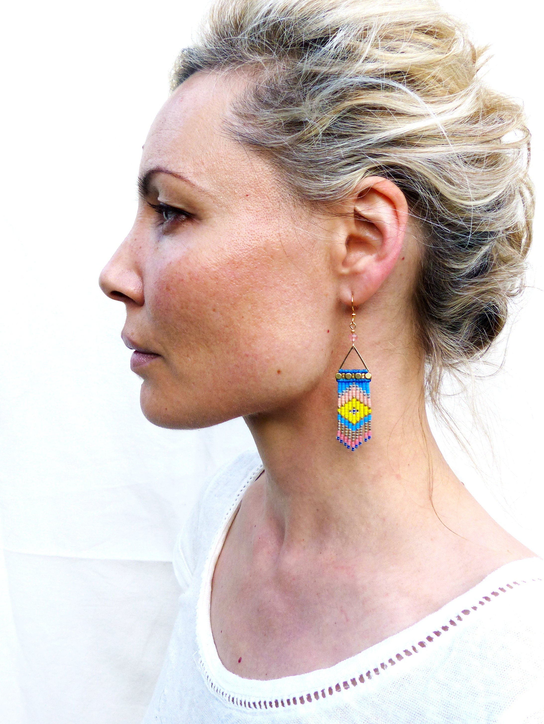 Peut inclure: Boucles d'oreilles géométriques en perles bleues, jaunes, roses et dorées. Les boucles d'oreilles sont rectangulaires avec une base triangulaire et sont suspendues à des crochets dorés. Le motif présente un losange jaune central.