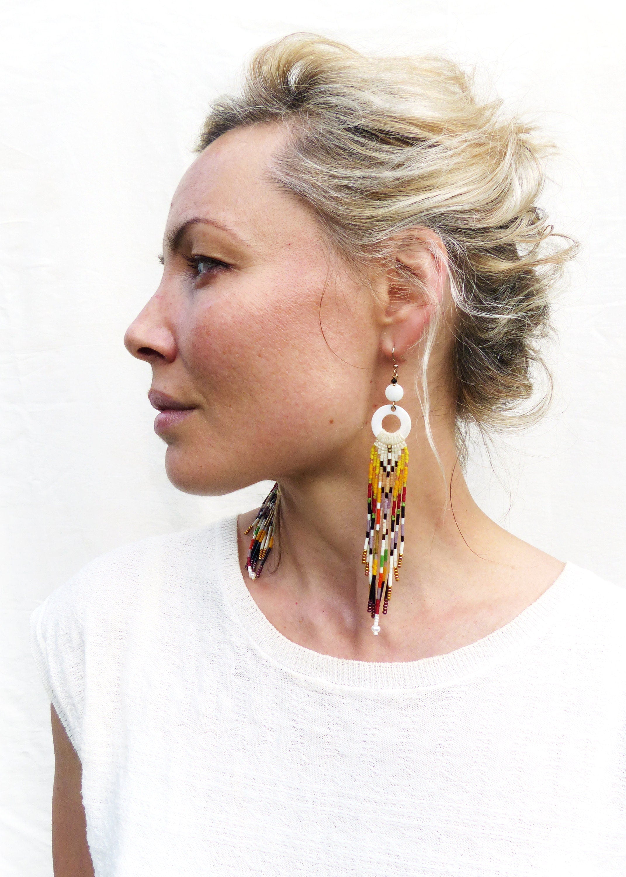 Peut inclure: Une paire de longues boucles d'oreilles en perles colorées avec un pendentif rond blanc. Les boucles d'oreilles sont faites avec une variété de perles dans des tons de rouge, orange, jaune, vert, bleu et violet. Les perles sont enfilées sur un fil fin et pendent en cascade.