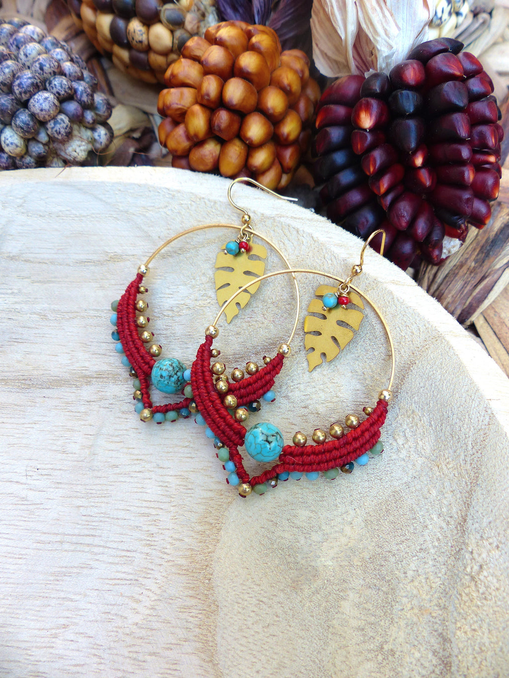 Peut inclure: Une paire de boucles d'oreilles créoles dorées avec des accents de perles rouges et turquoise. Les boucles d'oreilles présentent un charme en forme de feuille dorée et sont suspendues sur une surface en bois.