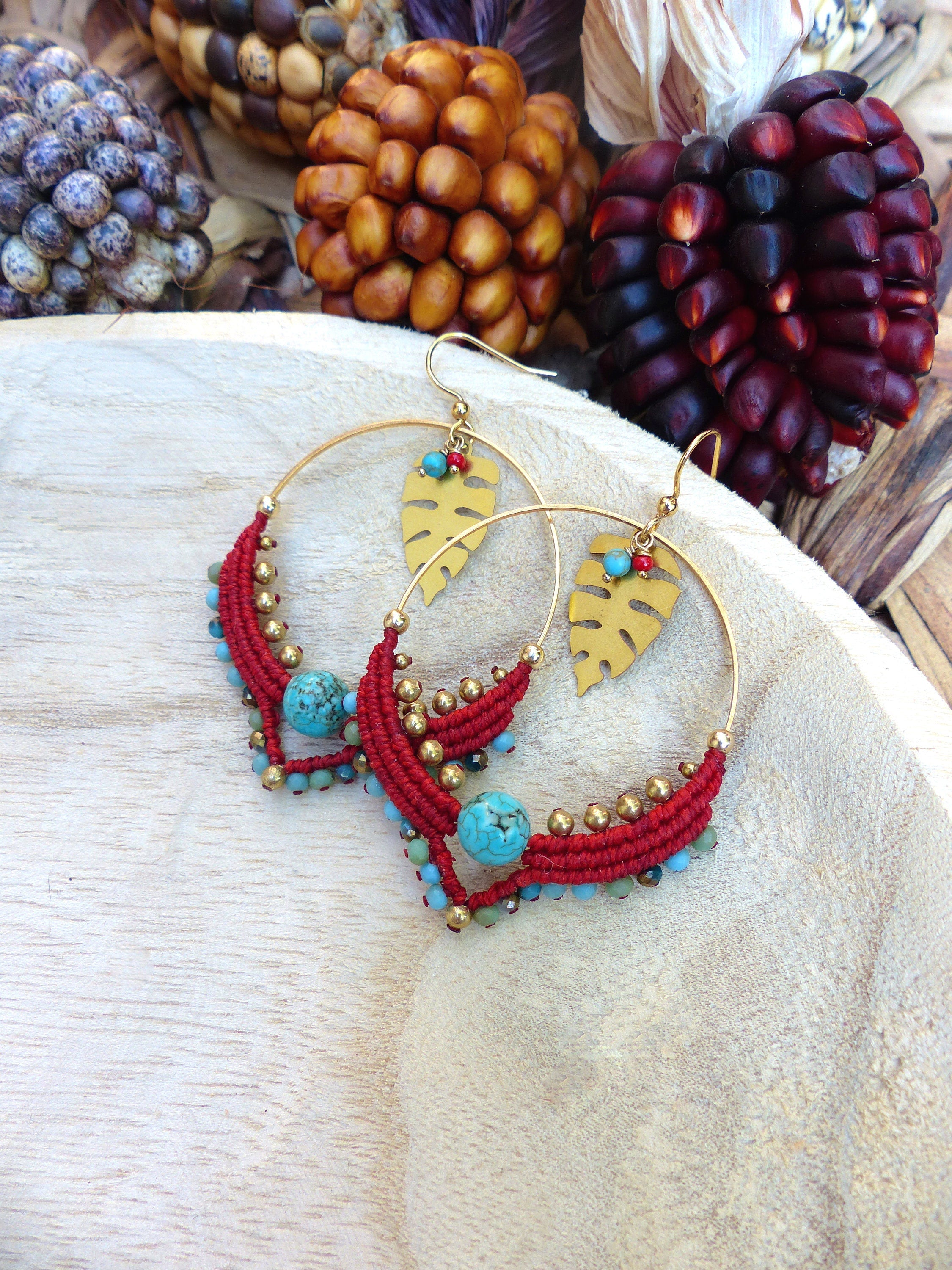 Peut inclure: Une paire de boucles d'oreilles créoles dorées avec des accents de perles rouges et turquoise. Les boucles d'oreilles présentent un charme en forme de feuille dorée et sont suspendues sur une surface en bois.