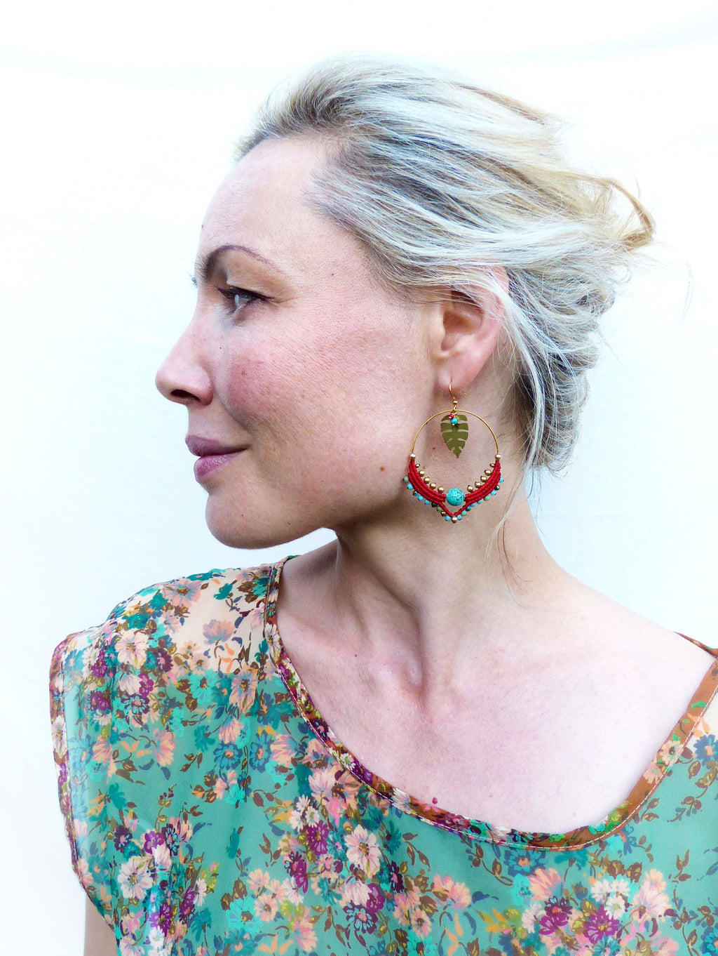Peut inclure: Une femme aux cheveux blonds portant un haut à imprimé floral vert et de grandes boucles d'oreilles en or avec des perles rouges et turquoise.