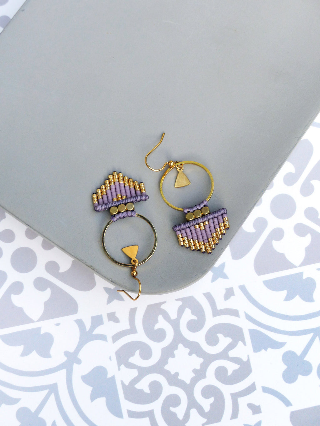 Peut inclure: Une paire de boucles d'oreilles en perles violettes et dorées avec un anneau et un charme triangulaire. Les boucles d'oreilles sont sur une surface grise.