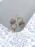 Peut inclure: Une paire de boucles d'oreilles en perles violettes et dorées avec un anneau et un charme triangulaire. Les boucles d'oreilles sont sur une surface grise.