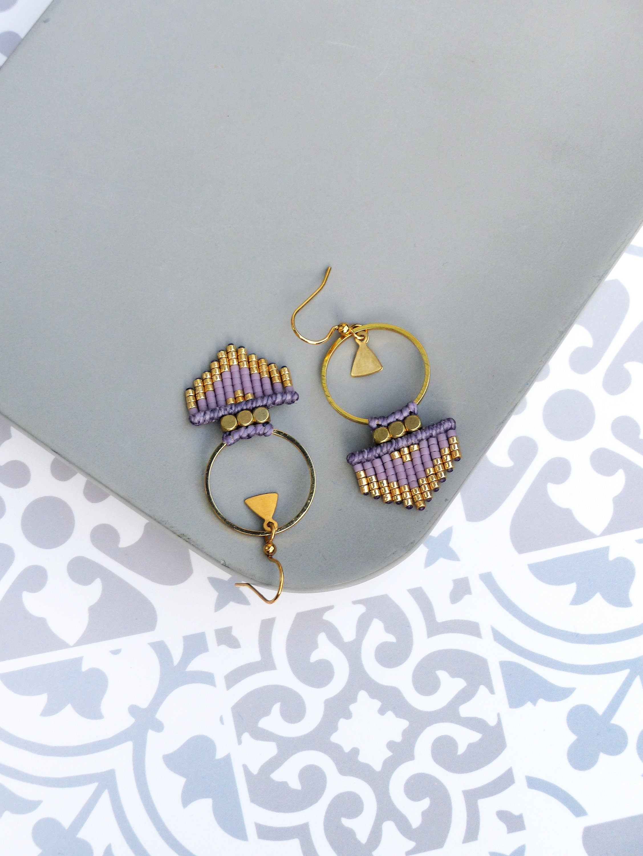 Peut inclure: Une paire de boucles d'oreilles en perles violettes et dorées avec un anneau et un charme triangulaire. Les boucles d'oreilles sont sur une surface grise.