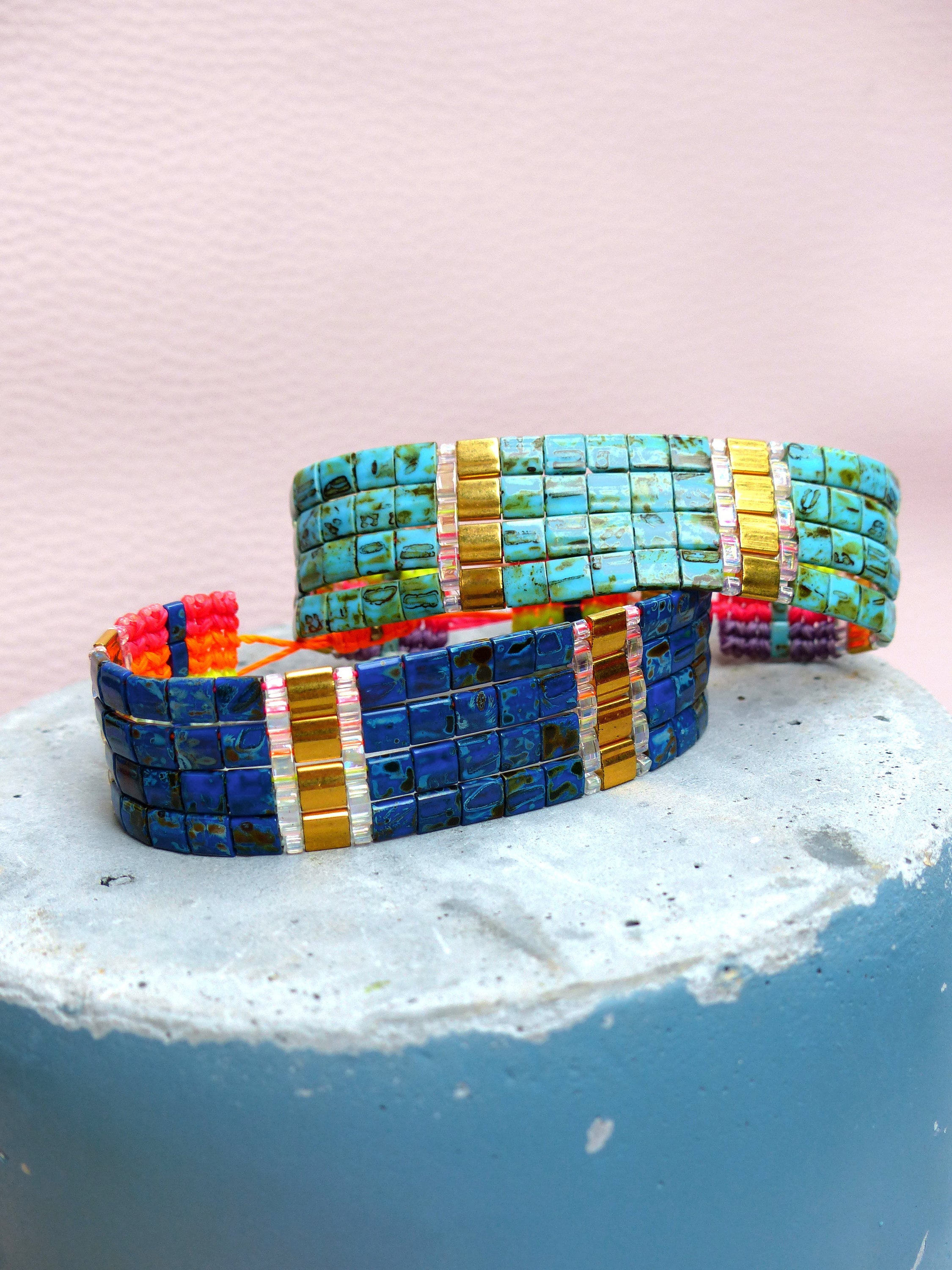 Peut inclure: Trois bracelets en perles colorées avec des accents dorés. Les bracelets sont empilés les uns sur les autres et sont faits de petites perles carrées. Les perles sont dans des tons de bleu, turquoise et or.