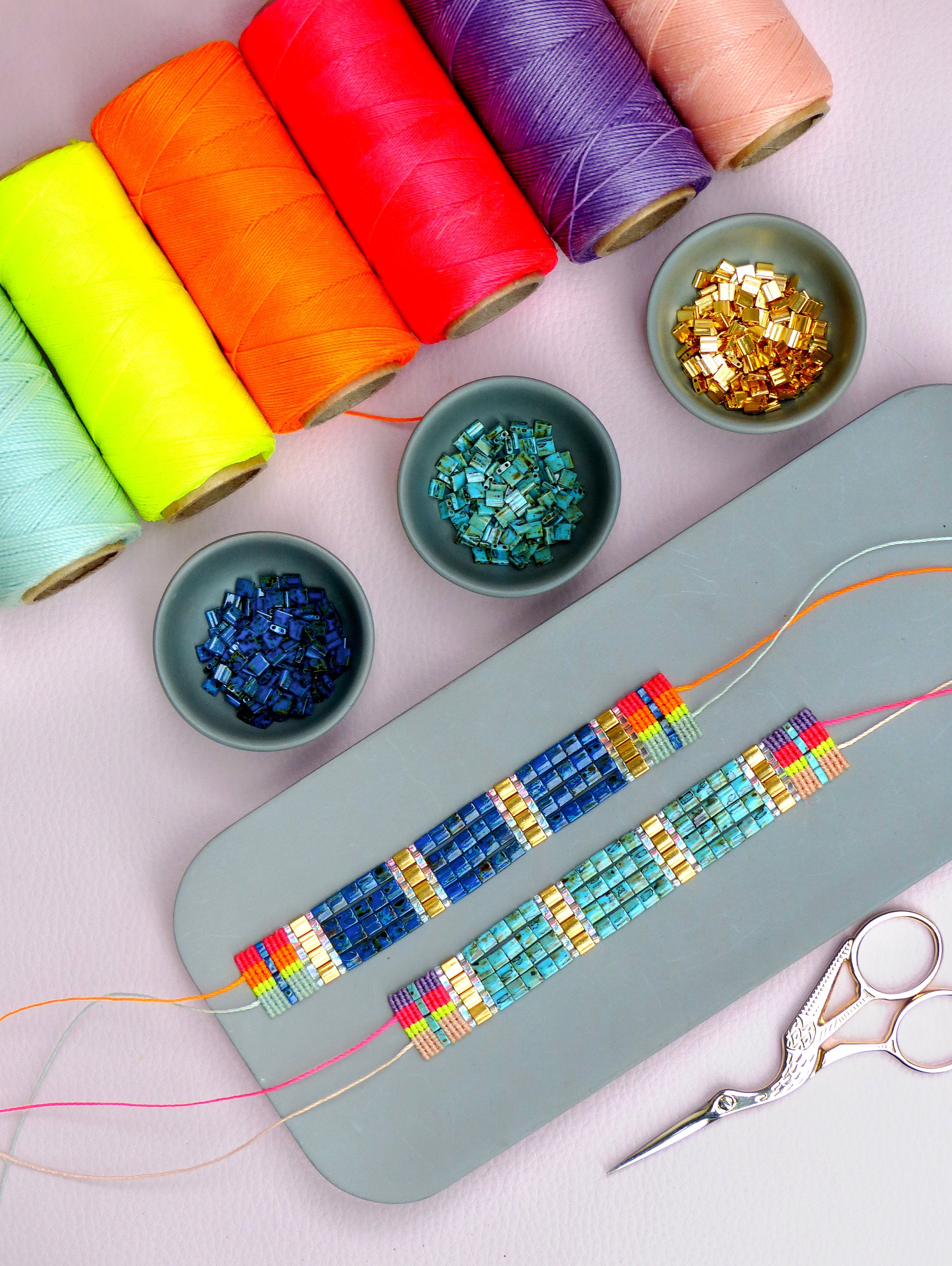 Peut inclure: Deux bracelets en perles avec des perles turquoise, bleues et dorées. Les bracelets sont sur une surface grise avec des bobines de fil coloré et de petits bols de perles.